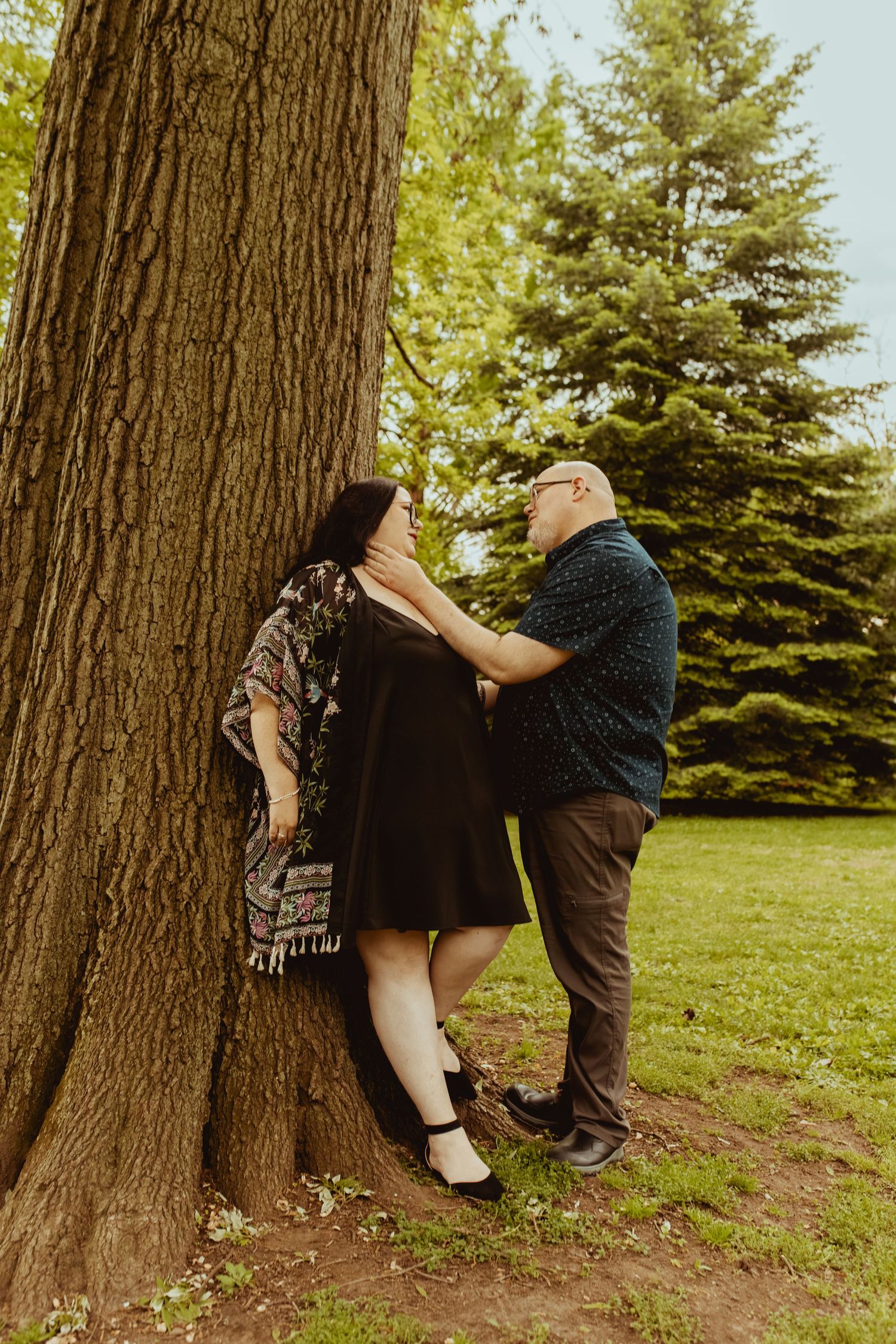 Couple sharing a romantic moment leaning against a large tree in a lush green park, woman in black dress.