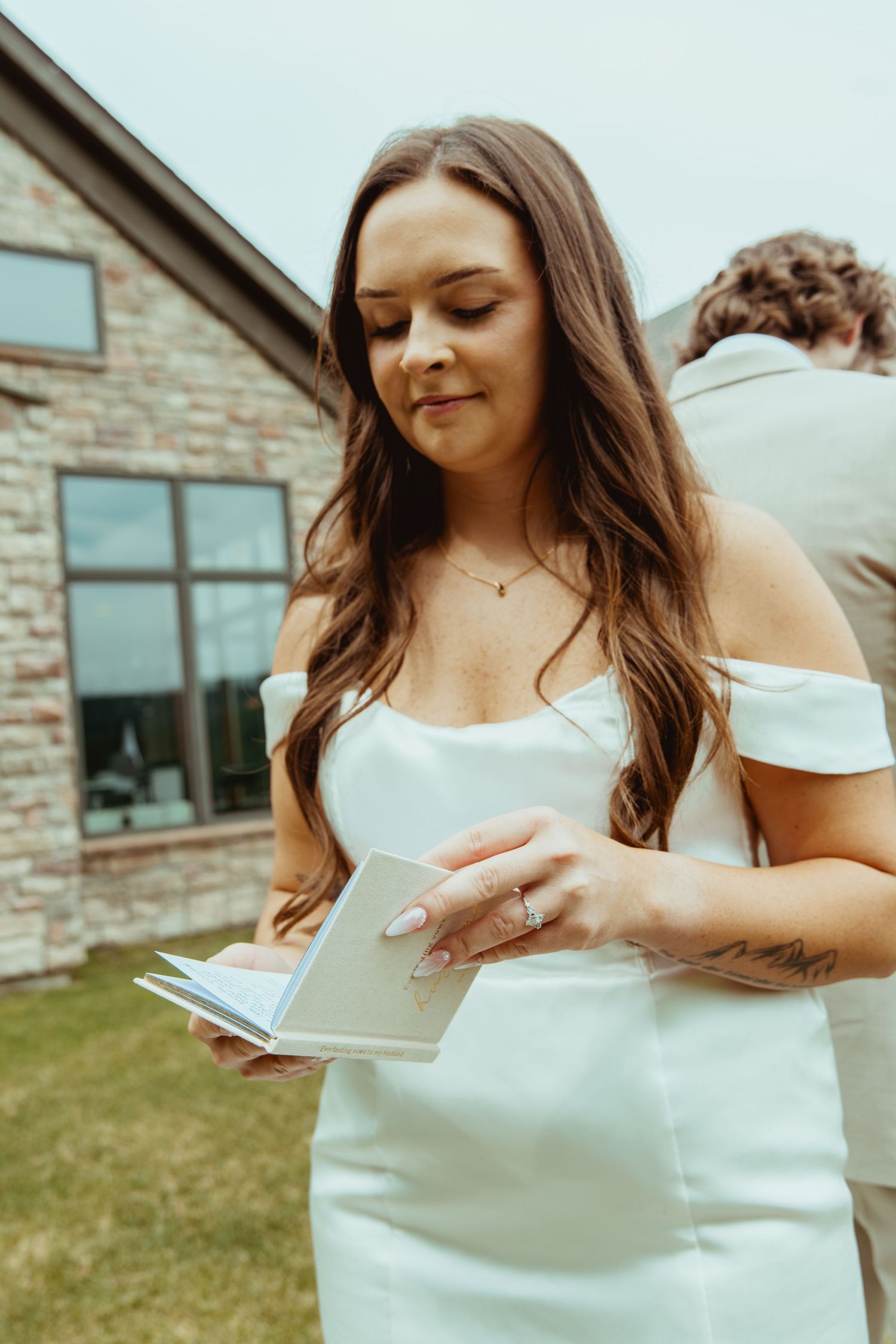 Brunette bride in off-shoulder white gown reads vows from booklet outside modern stone building on wedding day.