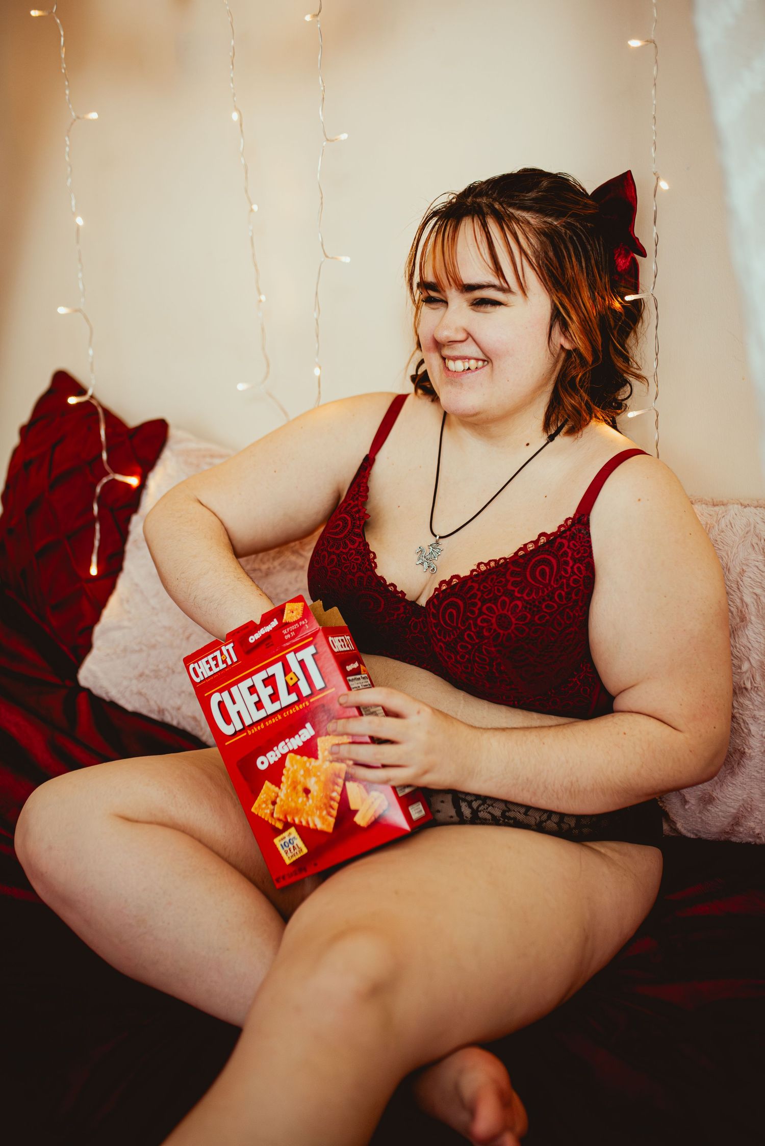 A person in red lingerie lounges on a couch holding a box of Cheez-It crackers in a softly lit room with fairy lights.