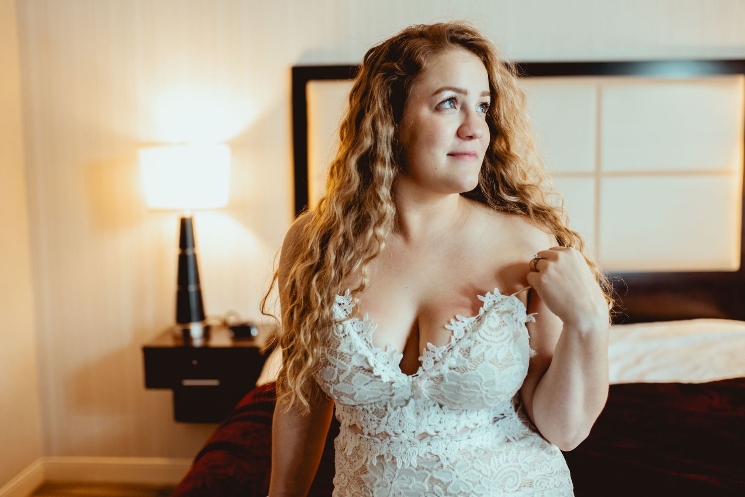 A bride in a white lace wedding dress sits on a bed in a warmly lit room during a bridal photoshoot.