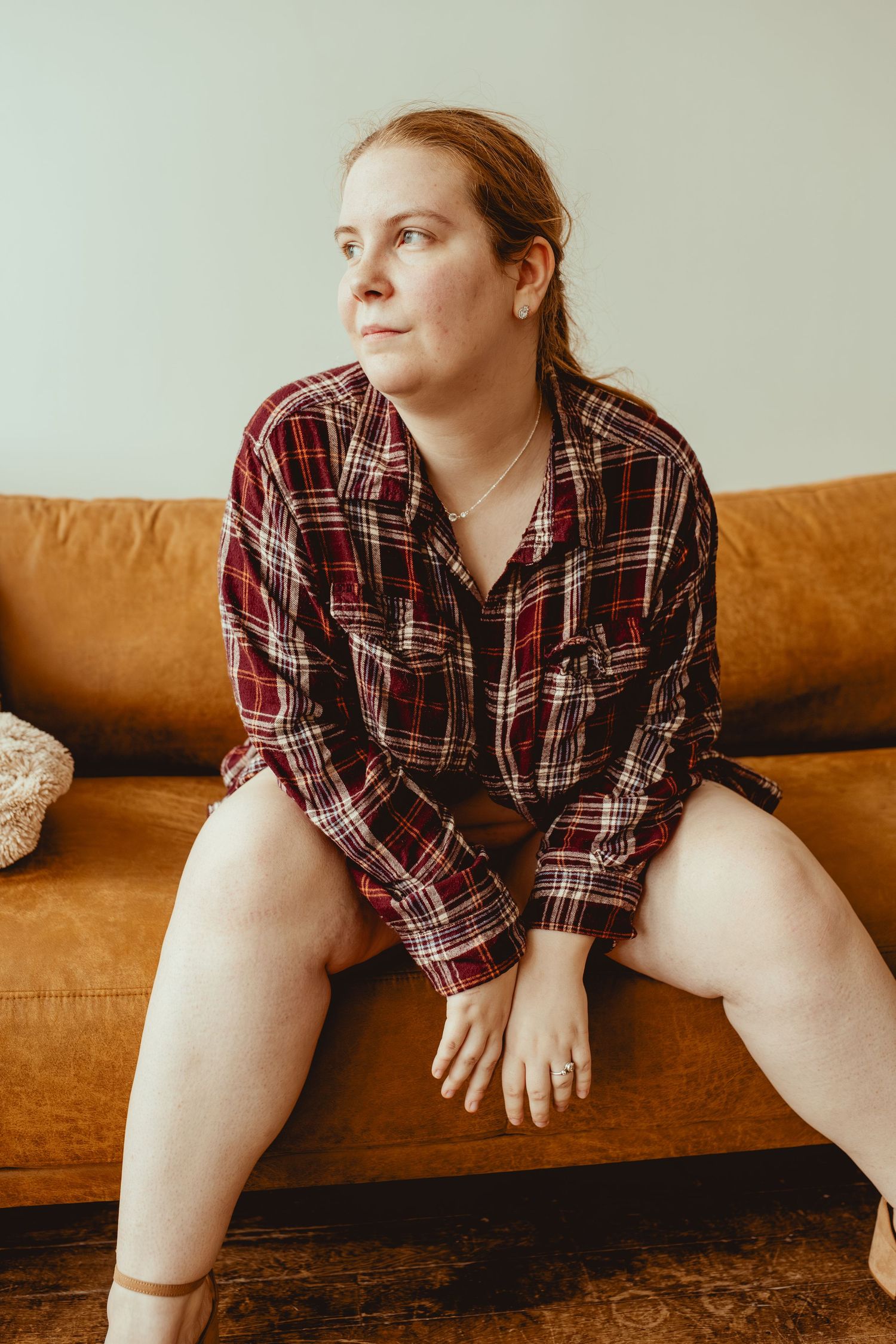 Person wearing plaid shirt sits casually on brown leather couch in warm sunlit room.