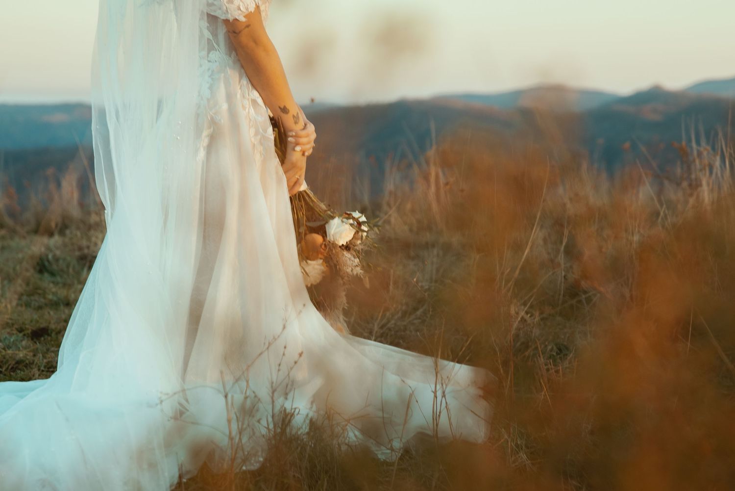 A bride in a flowing white dress stands against a scenic mountain backdrop during golden hour.