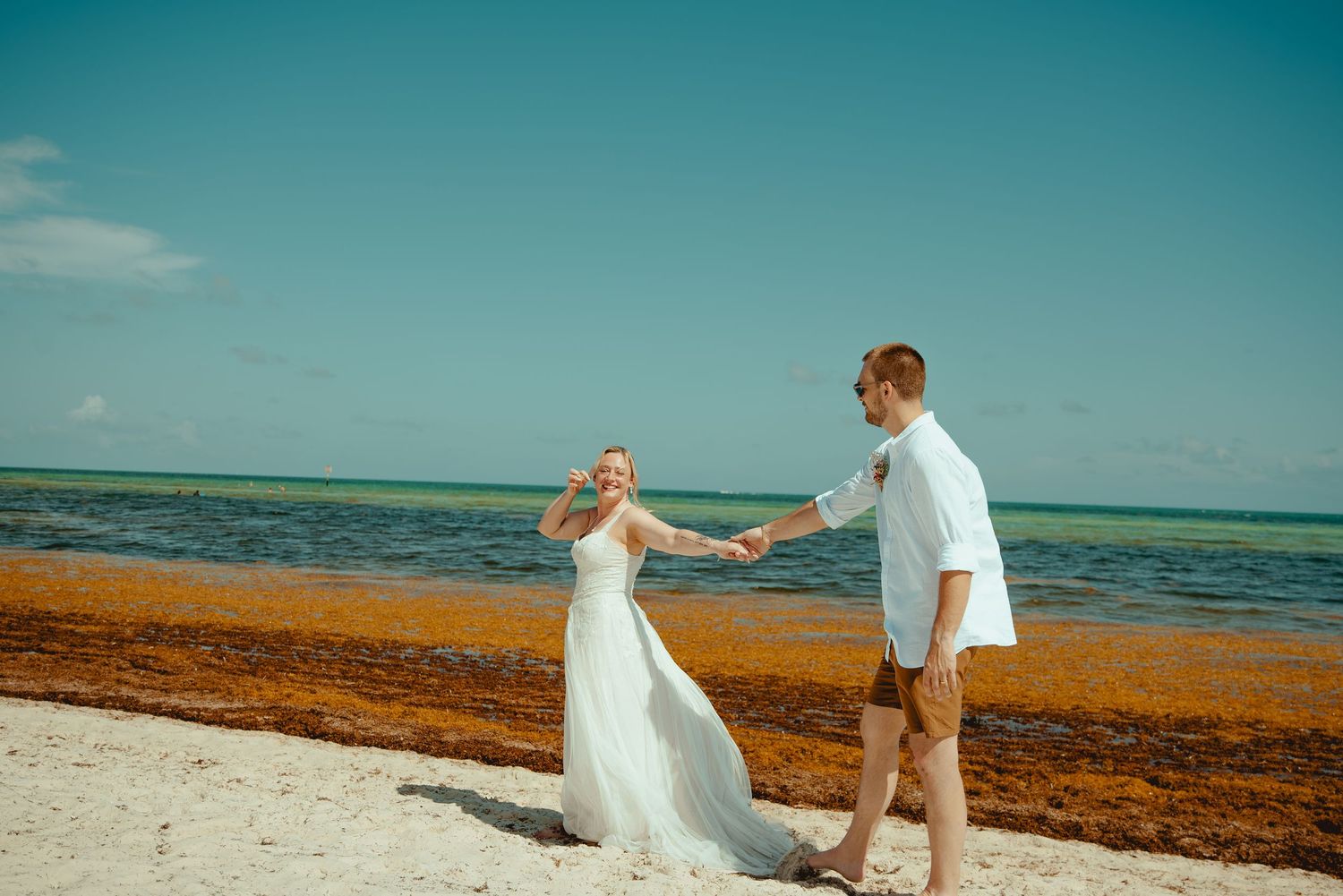 Bride and groom holding hands walking on a tropical beach with turquoise water and seaweed shore.