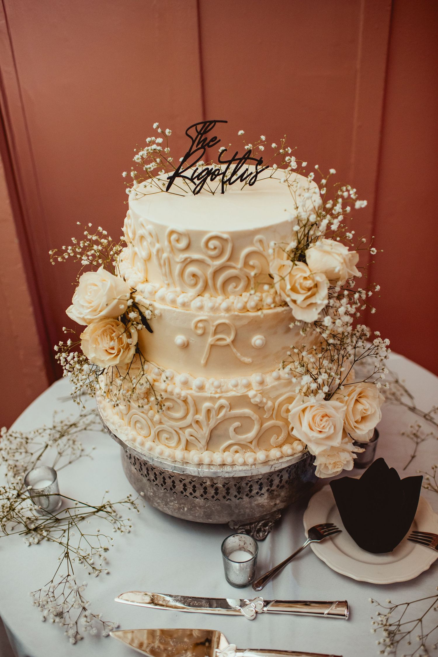Elegant white wedding cake with delicate piped swirls and fresh roses, displayed on a silver cake stand with baby's breath accents.