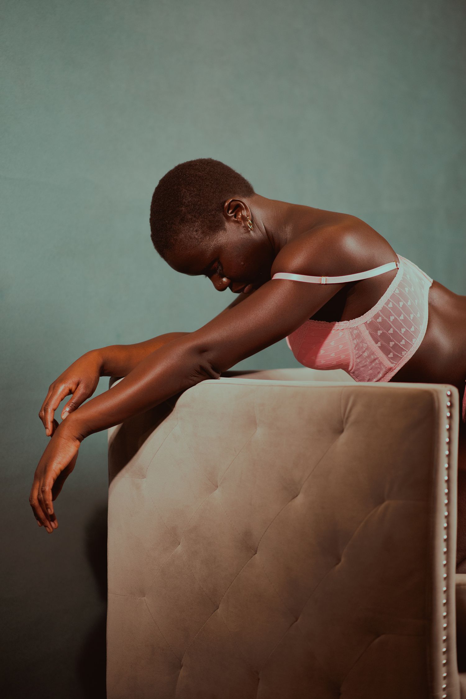 A Black woman in pink and white lingerie leans gracefully over a tufted beige headboard against a muted teal wall.