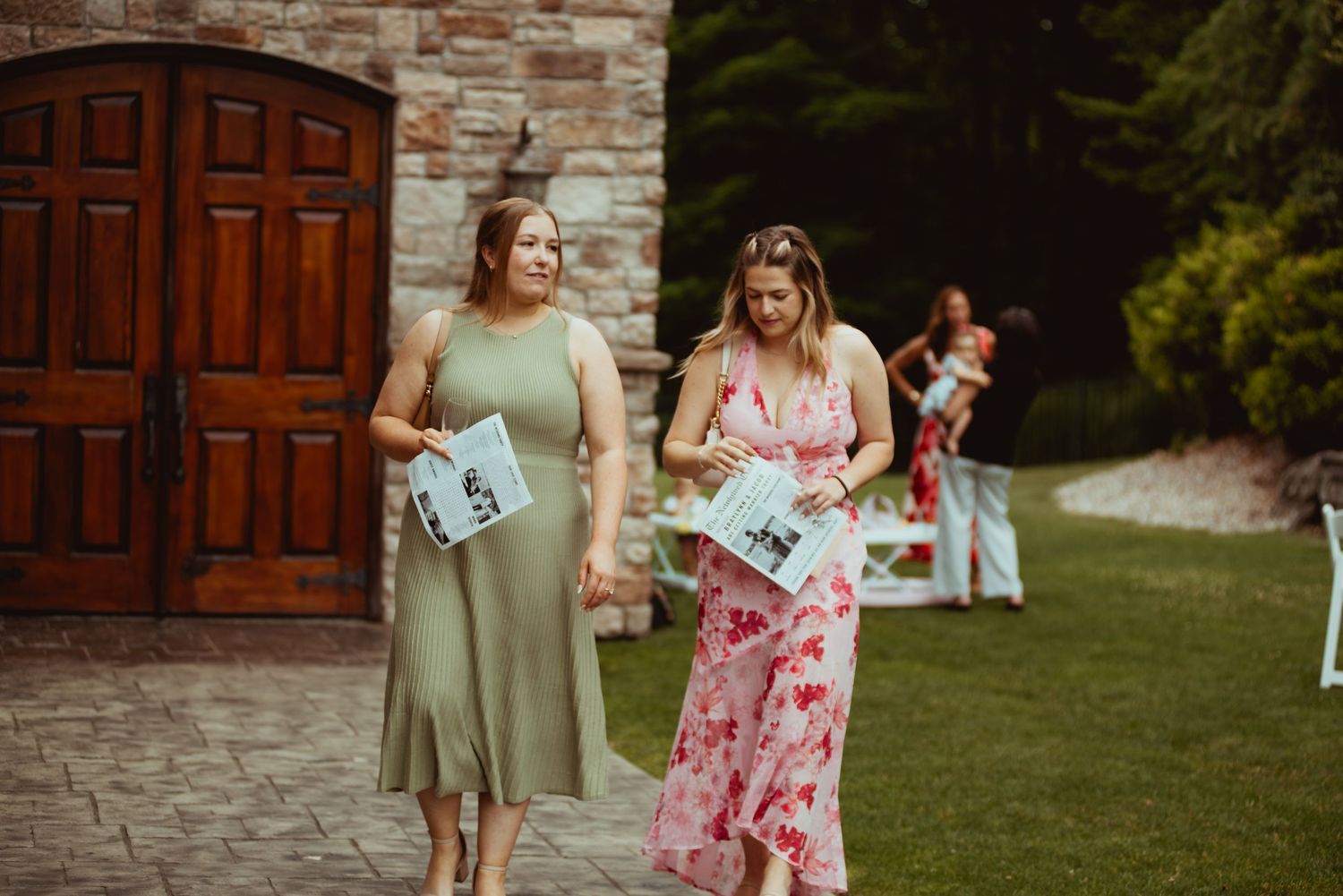 Two bridesmaids in green and floral dresses carrying wedding programs outside stone venue.