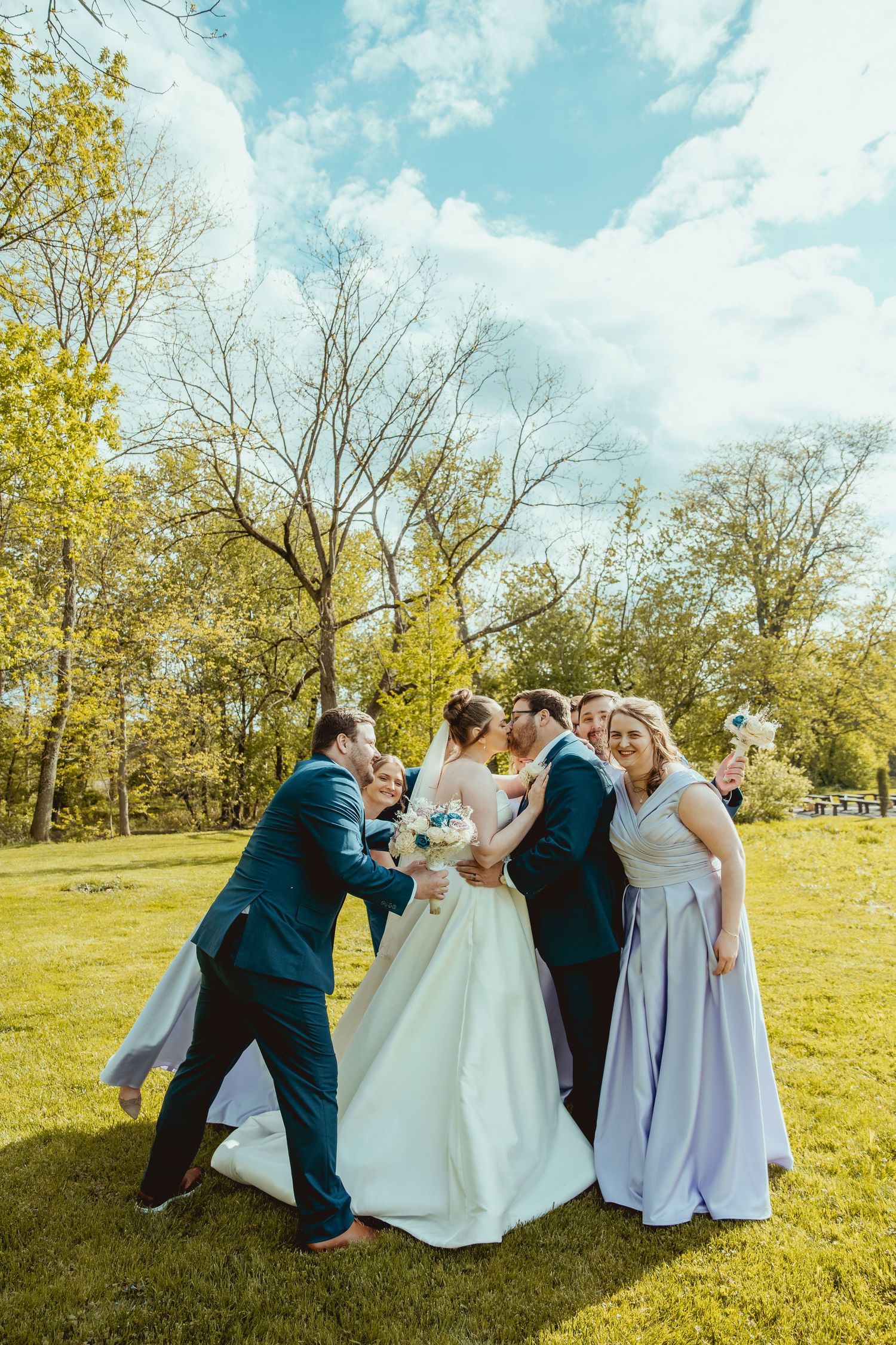 Bridal party celebrating outdoors as groomsmen kiss bride and bridesmaids cheer in sunny park.
