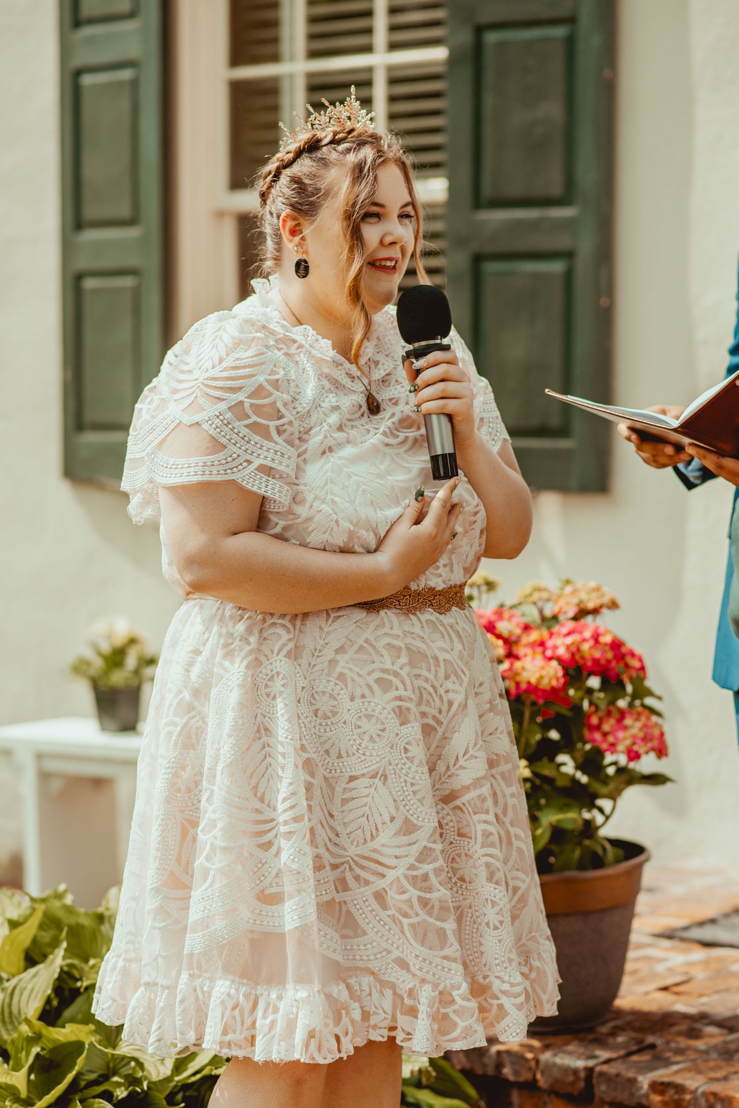 A person in a white lace dress holds a drink near a house with green shutters and potted flowers.
