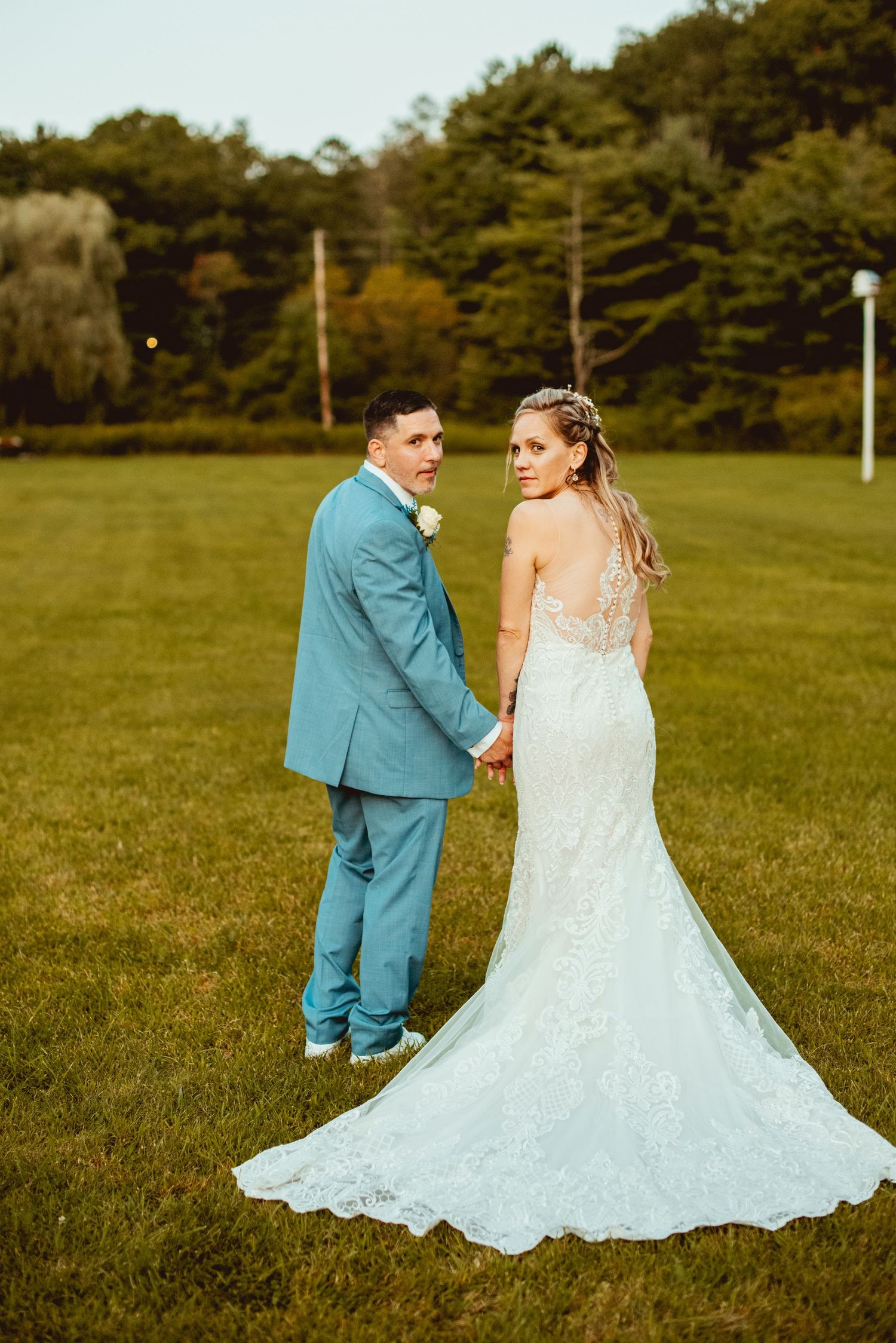 Wedding couple posing in grassy field with pine trees in background.