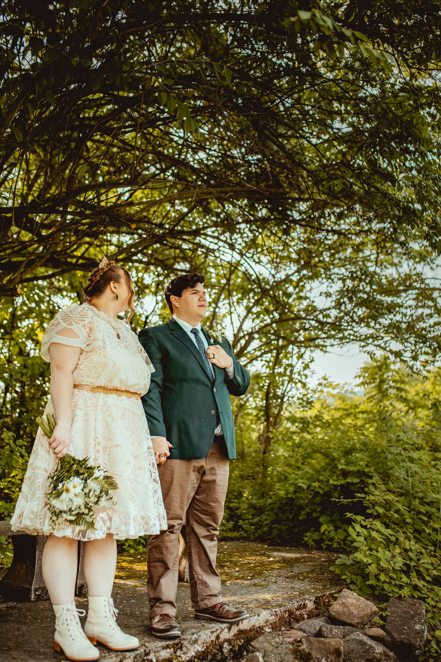 A couple in white dress and brown pants stand beneath leafy trees on a rocky forest path during a sunny day.