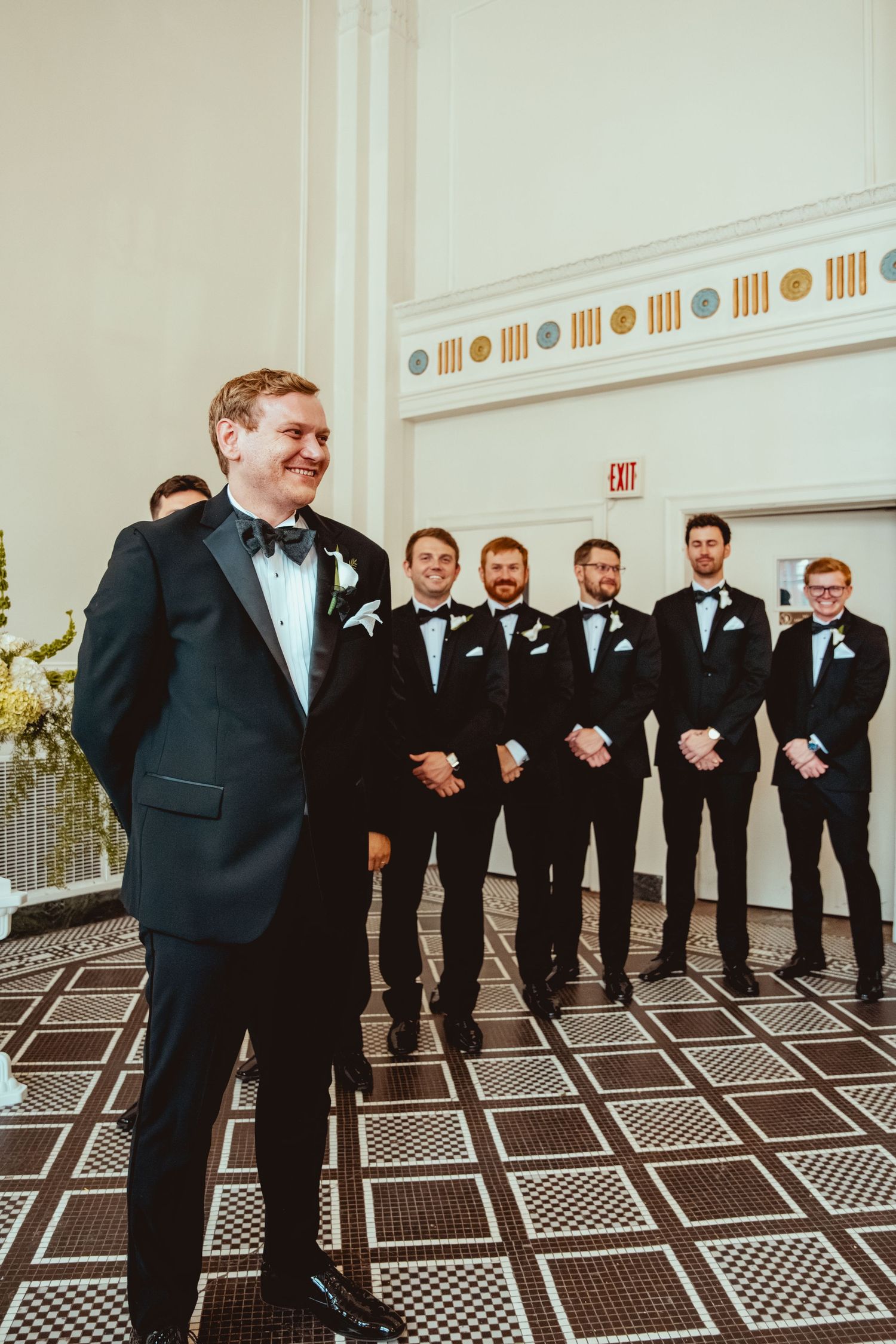 Groomsmen in black tuxedos laughing together on a geometric patterned floor.