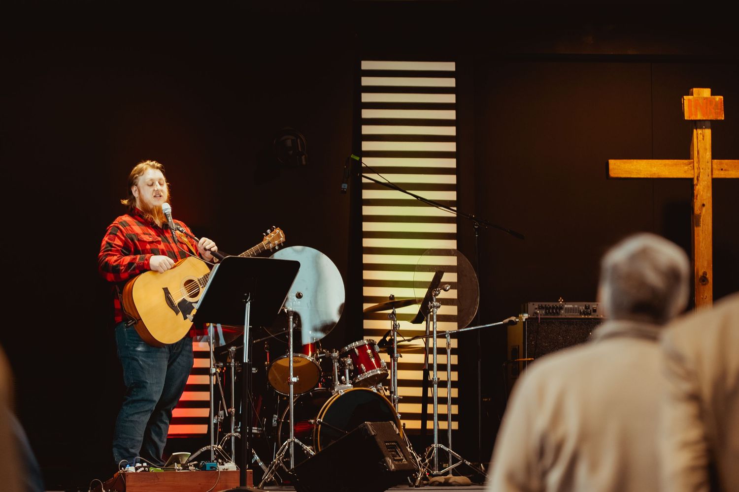 Musicians perform on stage in a church setting with acoustic guitar and drums visible in warm lighting.