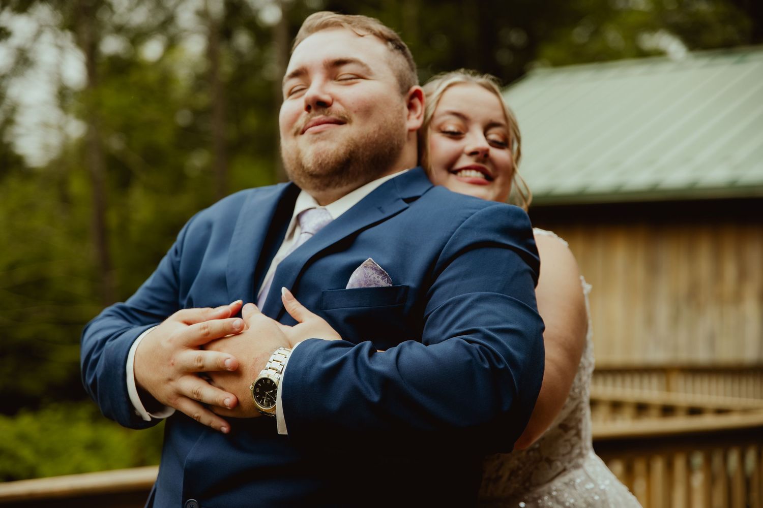 Bride hugs groom from behind as both smile with eyes closed in a joyful wedding portrait outdoors.