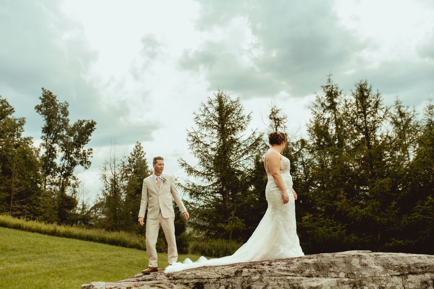 Bride and groom stand apart on rocky outcrop surrounded by lush green trees under cloudy sky during outdoor wedding.