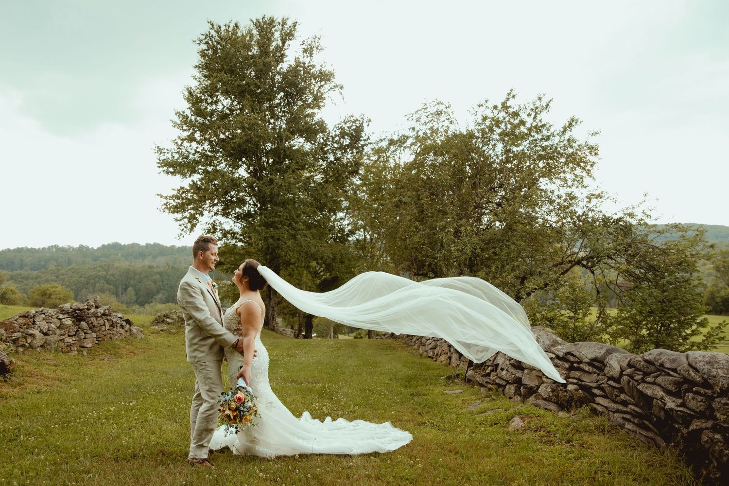 Bride and groom share a romantic moment outdoors as her veil flows in the wind beside a rustic stone wall.