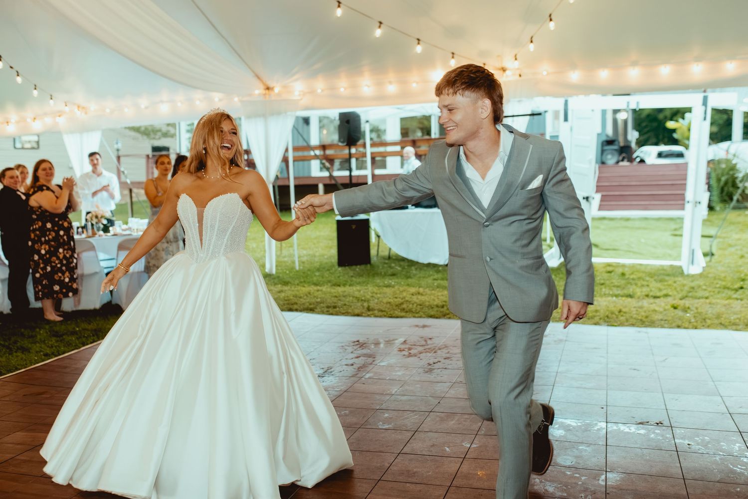Bride in white gown and groom in gray suit share first dance under a tented outdoor wedding reception.