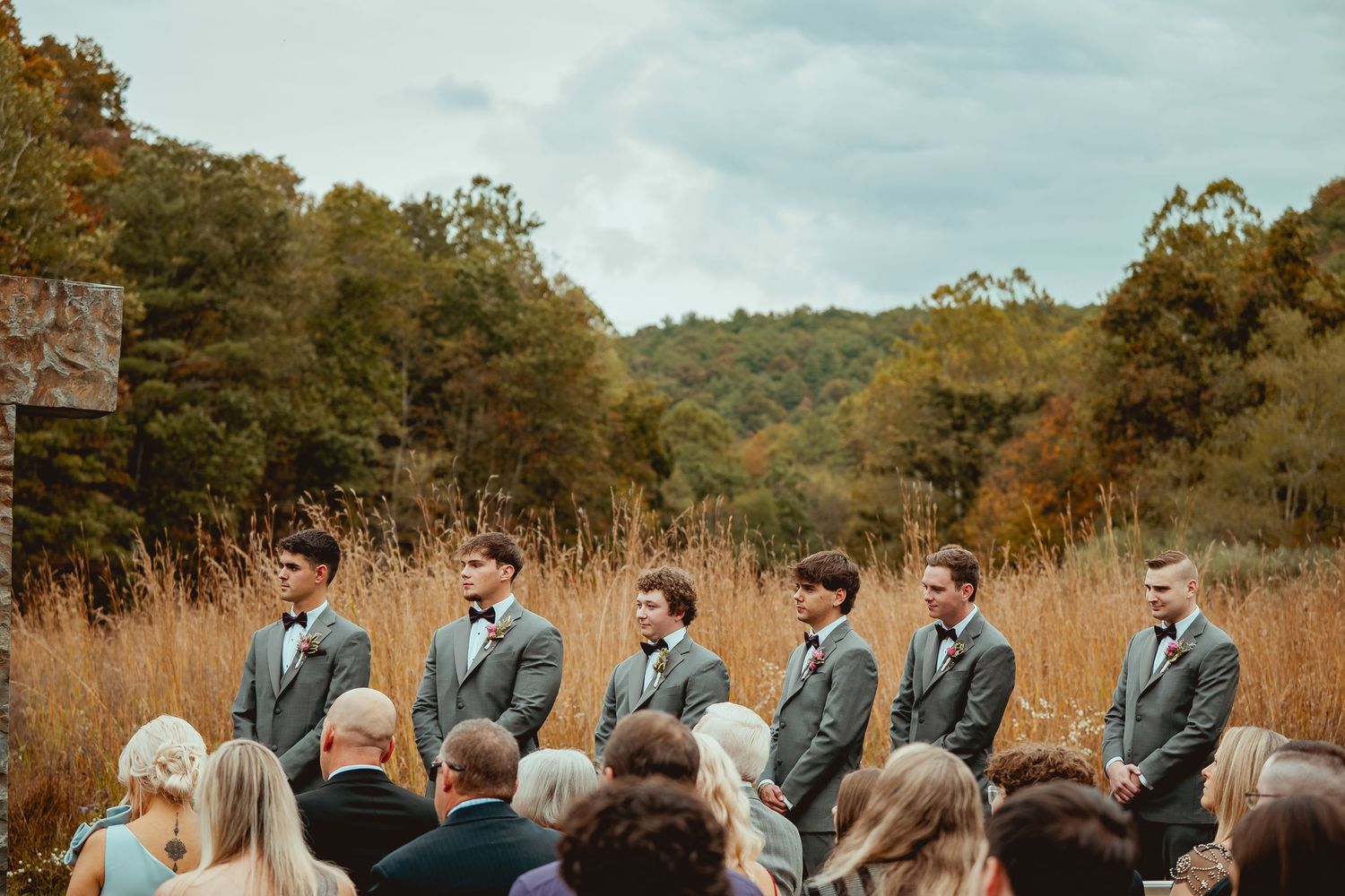 Wedding ceremony guests seated in white chairs on a grassy hill surrounded by autumn foliage.