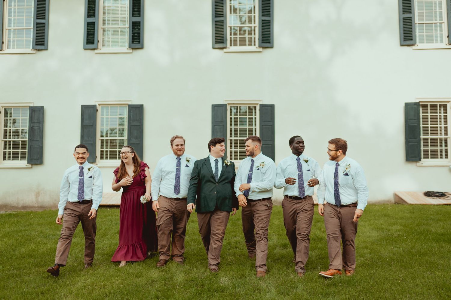 Wedding party members stand in a line on grass outside a white building with windows.
