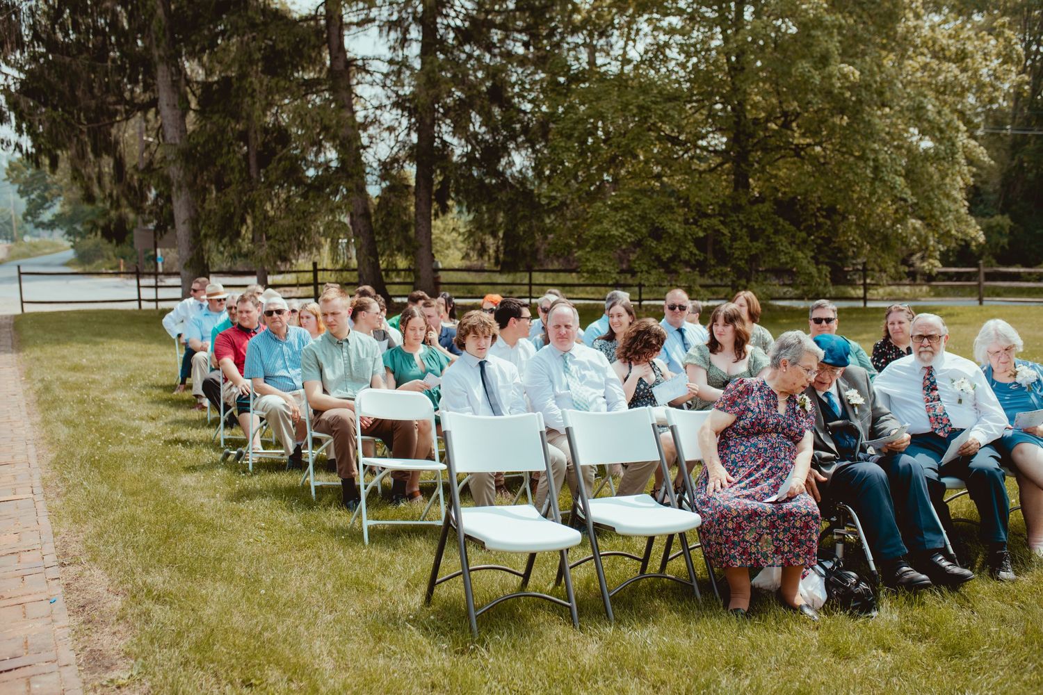Guests seated in white chairs on a lawn for an outdoor wedding ceremony with trees in the background.