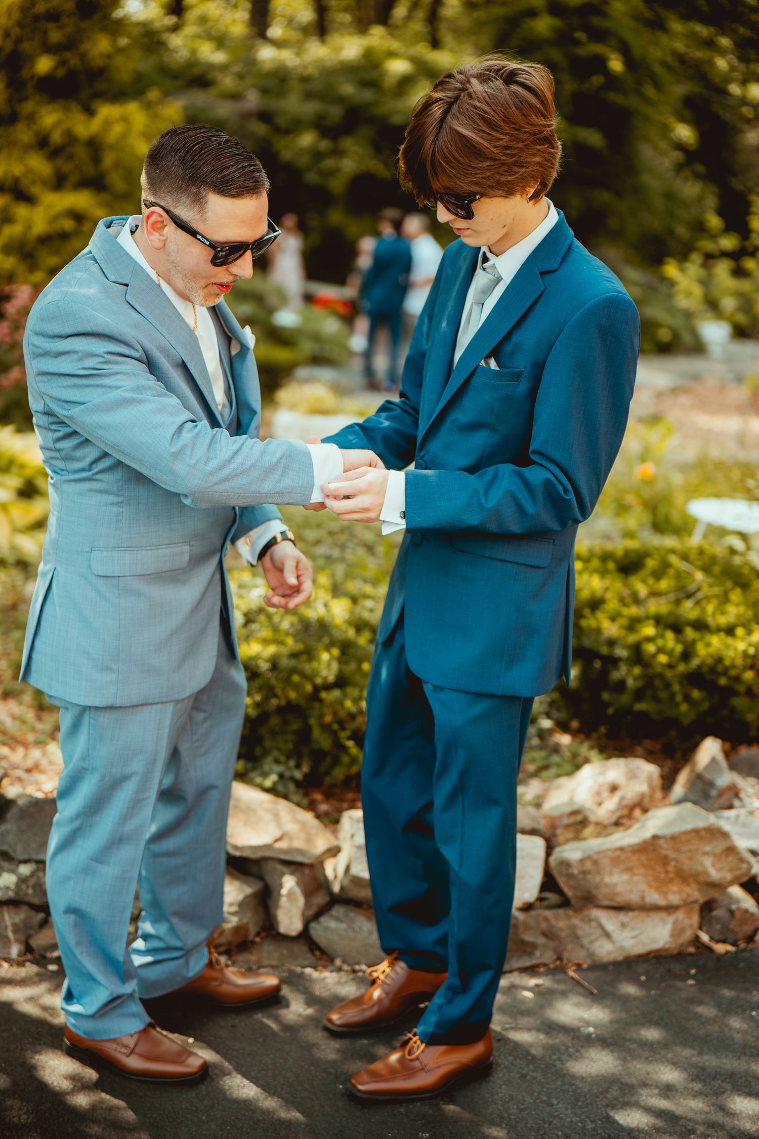 Two people in coordinating blue suits examine their shoes while standing outdoors on a stone path.