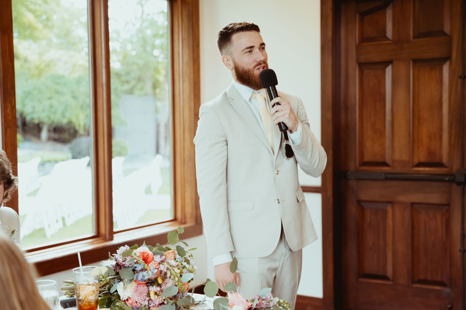 Bearded groom in beige suit delivers speech at wedding reception holding a microphone.