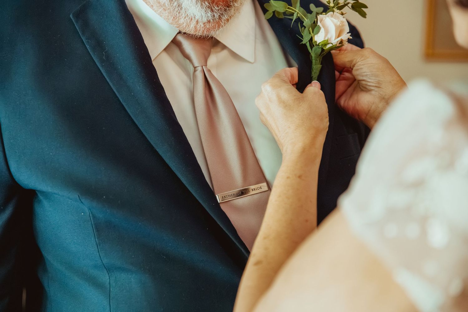 A bride pins a floral boutonniere on a groom wearing a navy suit and blush pink tie at a wedding.