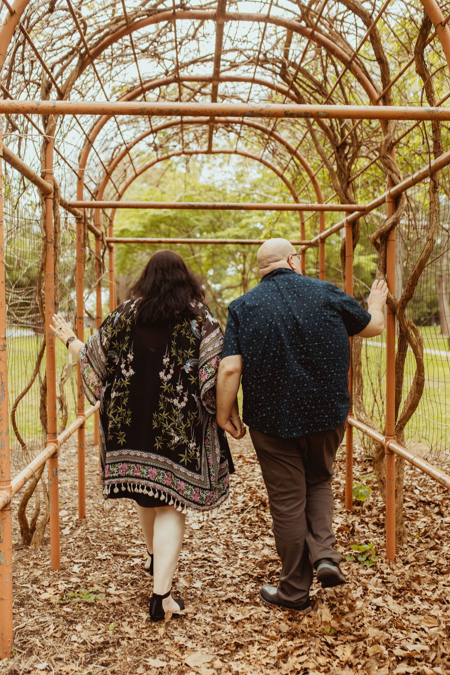 Couple walking hand in hand through a rustic wooden arbor tunnel covered in vines.