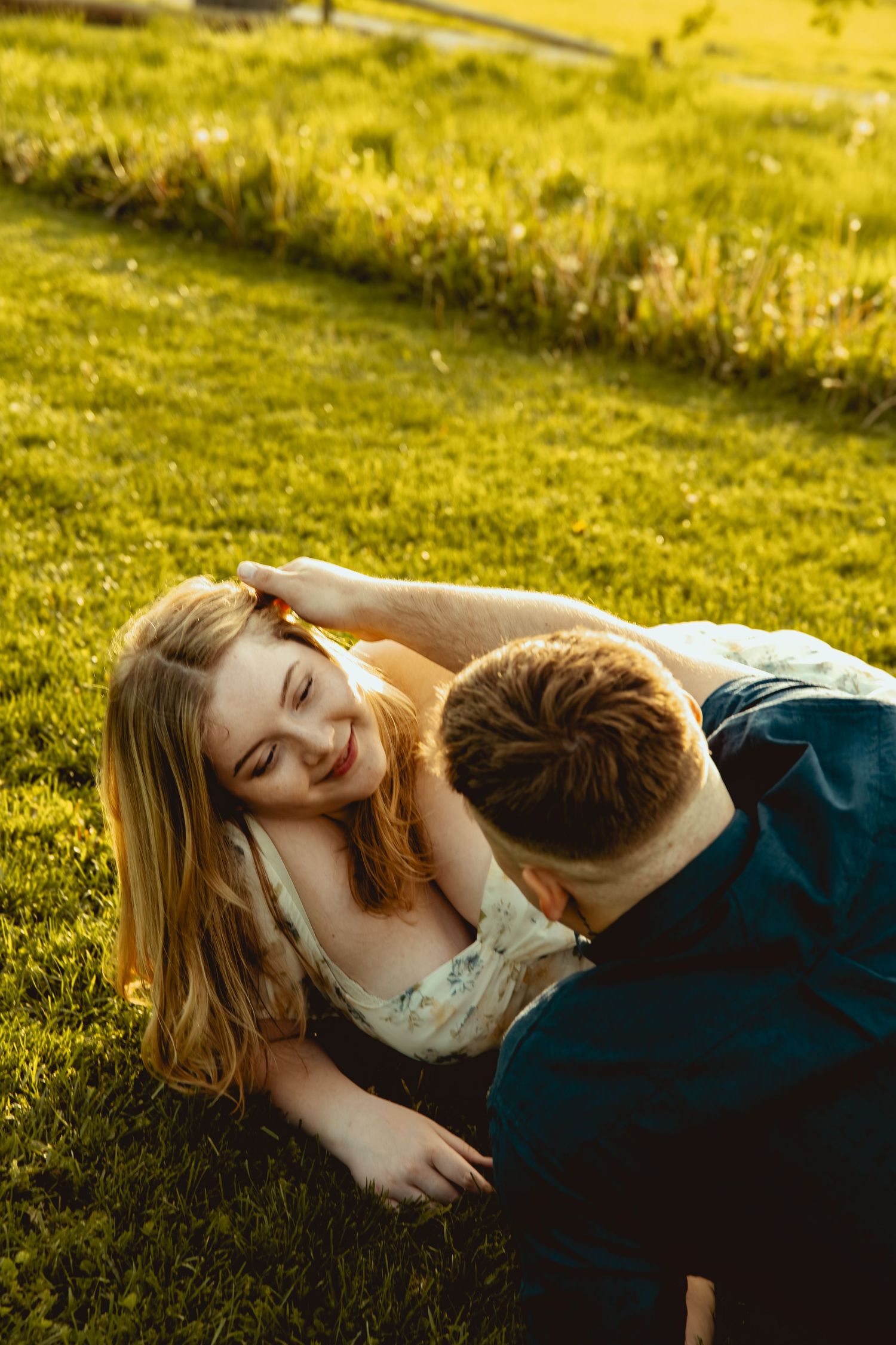 Couple lying on green grass in golden sunlight, woman smiling as man leans over her in romantic outdoor setting.