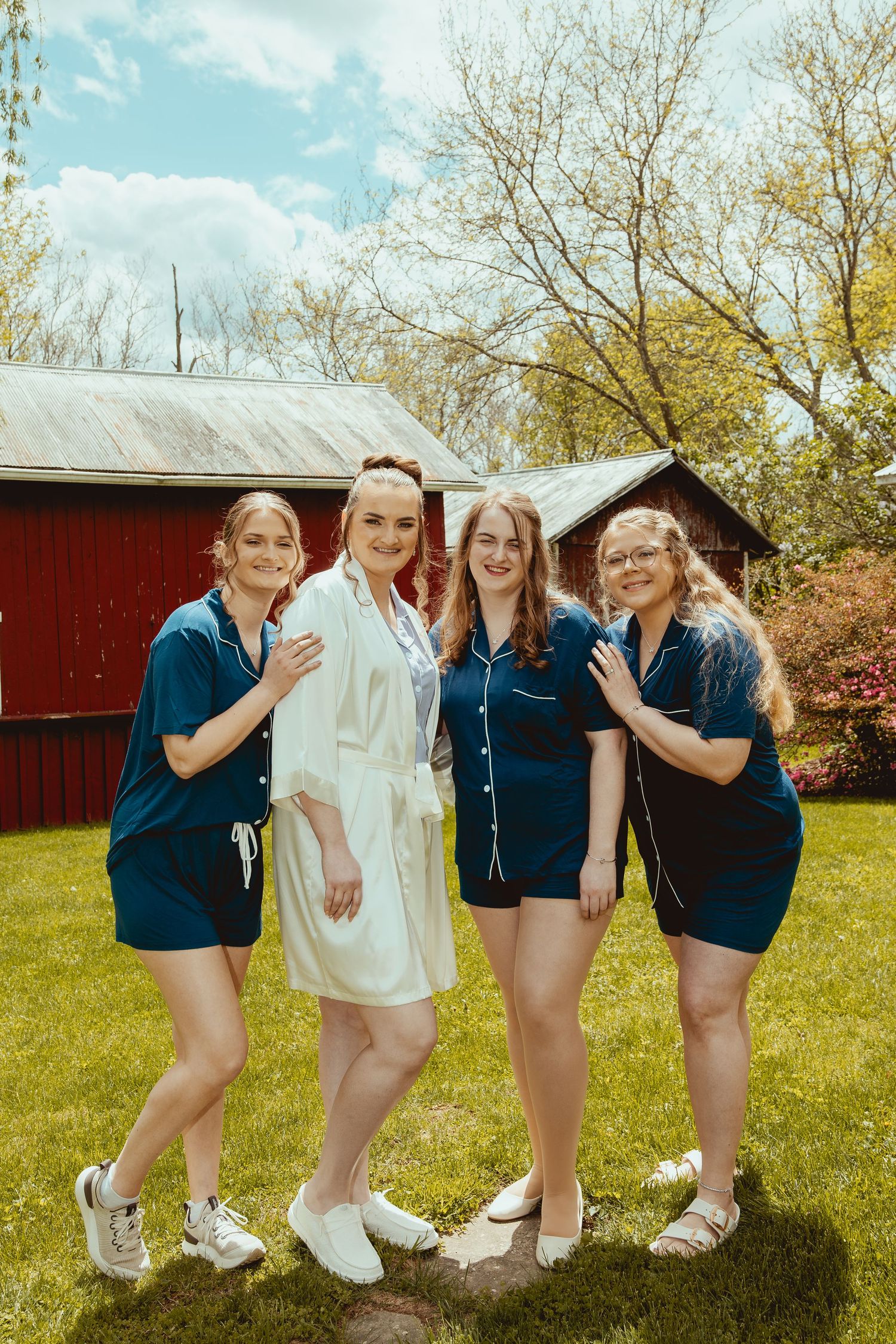 Bride in white robe posing with three bridesmaids in teal pajamas outdoors near red barn on spring day.