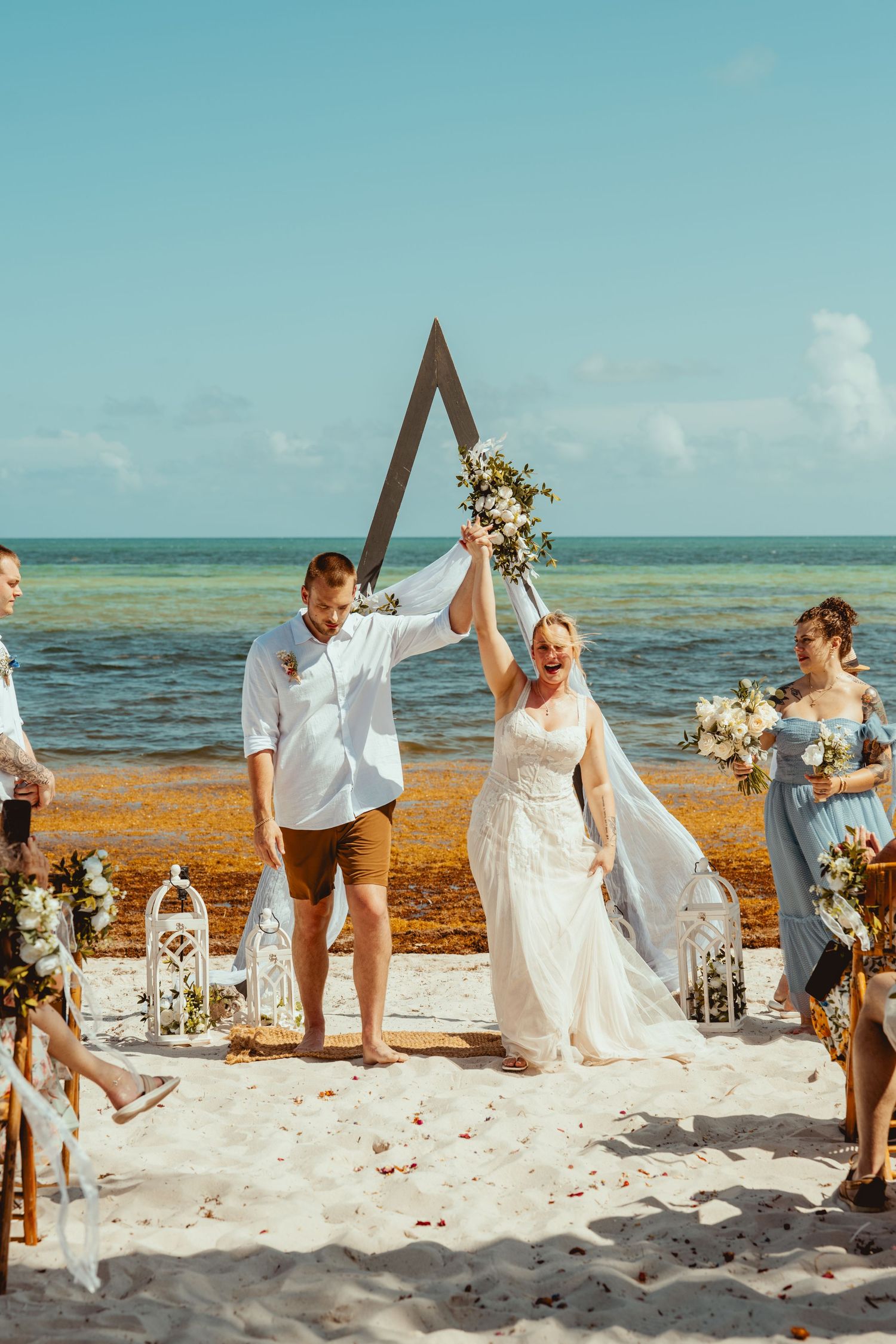 Bride and groom raise joined hands in celebration after their beachside wedding ceremony under a triangle arch by the turquoise sea.