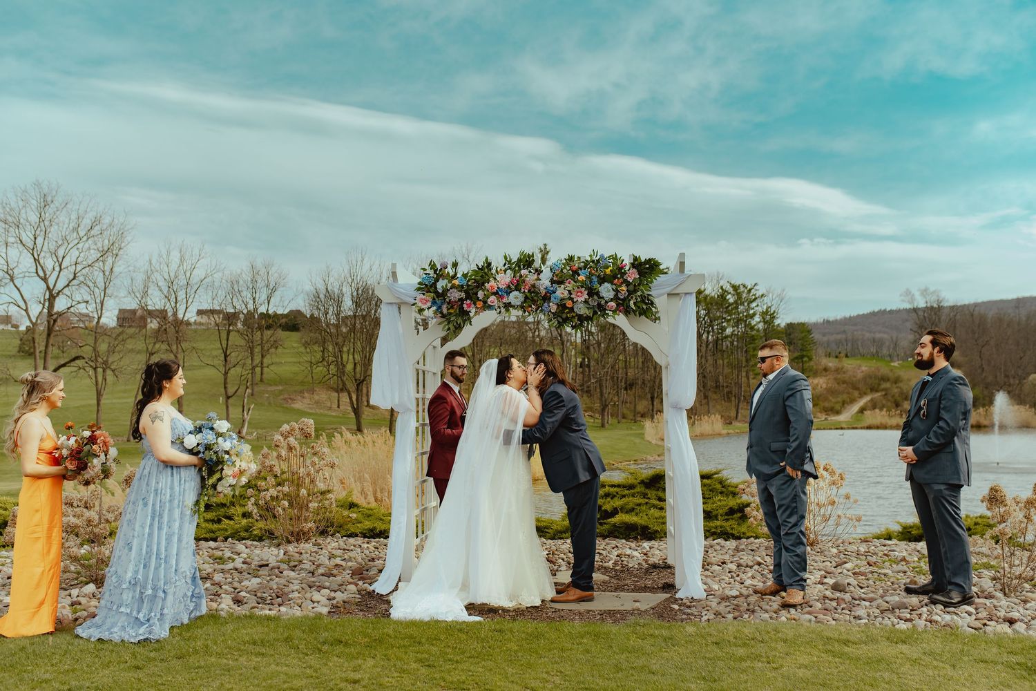 Outdoor wedding ceremony with couple kissing under floral arch by a lake, surrounded by wedding party in colorful attire.