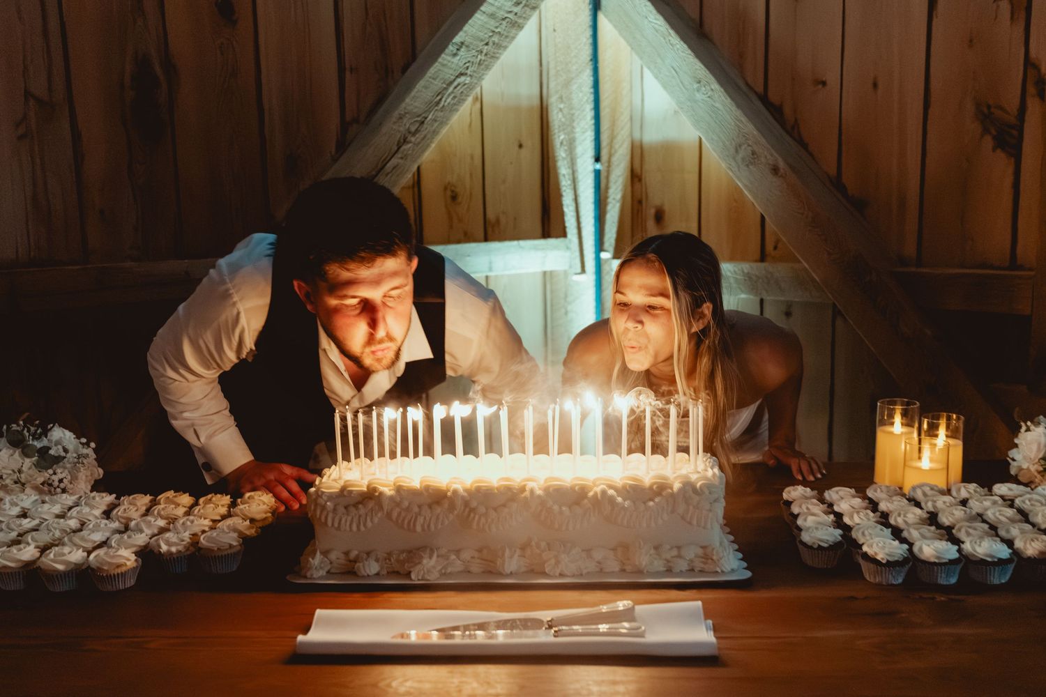 Couple blowing out candles on a large wedding cake surrounded by cupcakes in a warmly lit rustic venue.