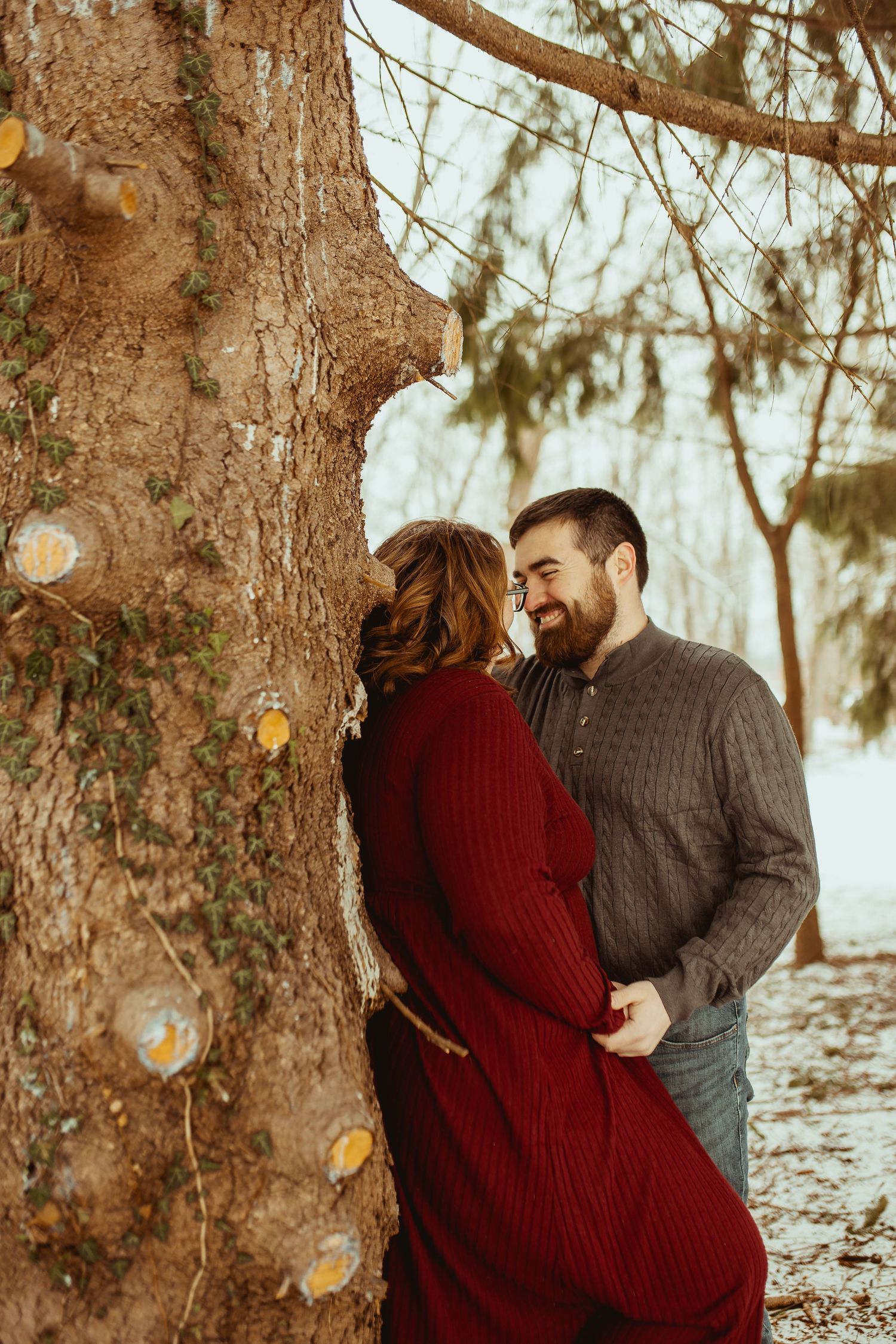 A couple shares a romantic moment in a snowy winter forest setting with sunlight filtering through pine trees.