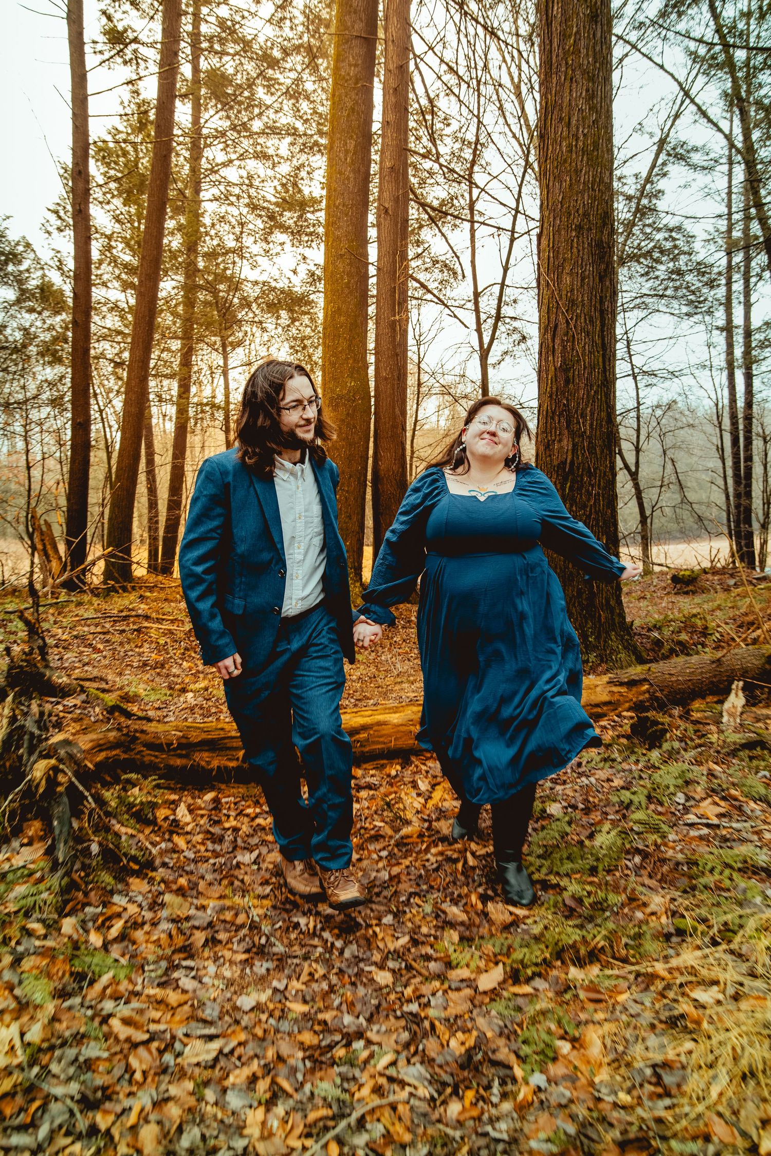 A couple wearing matching blue outfits walks through an autumn forest with golden sunlight filtering through trees.