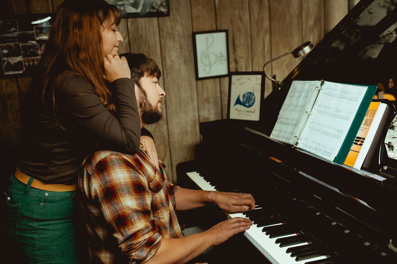 A person in a brown plaid shirt plays a black upright piano in a wood-paneled room.