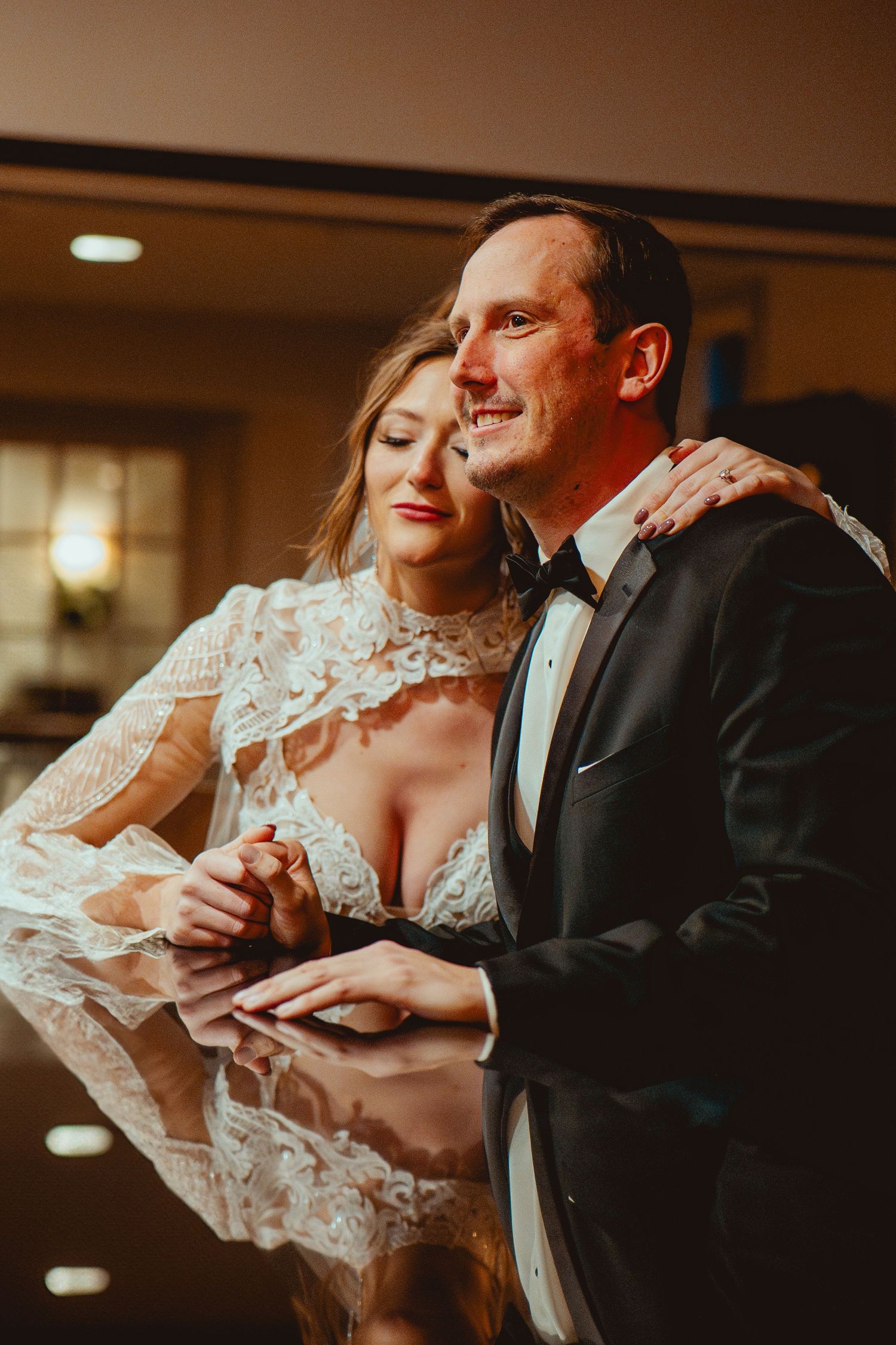Romantic wedding couple embraces in warm lighting with bride in lace dress and groom in tuxedo leaning on reflective surface.