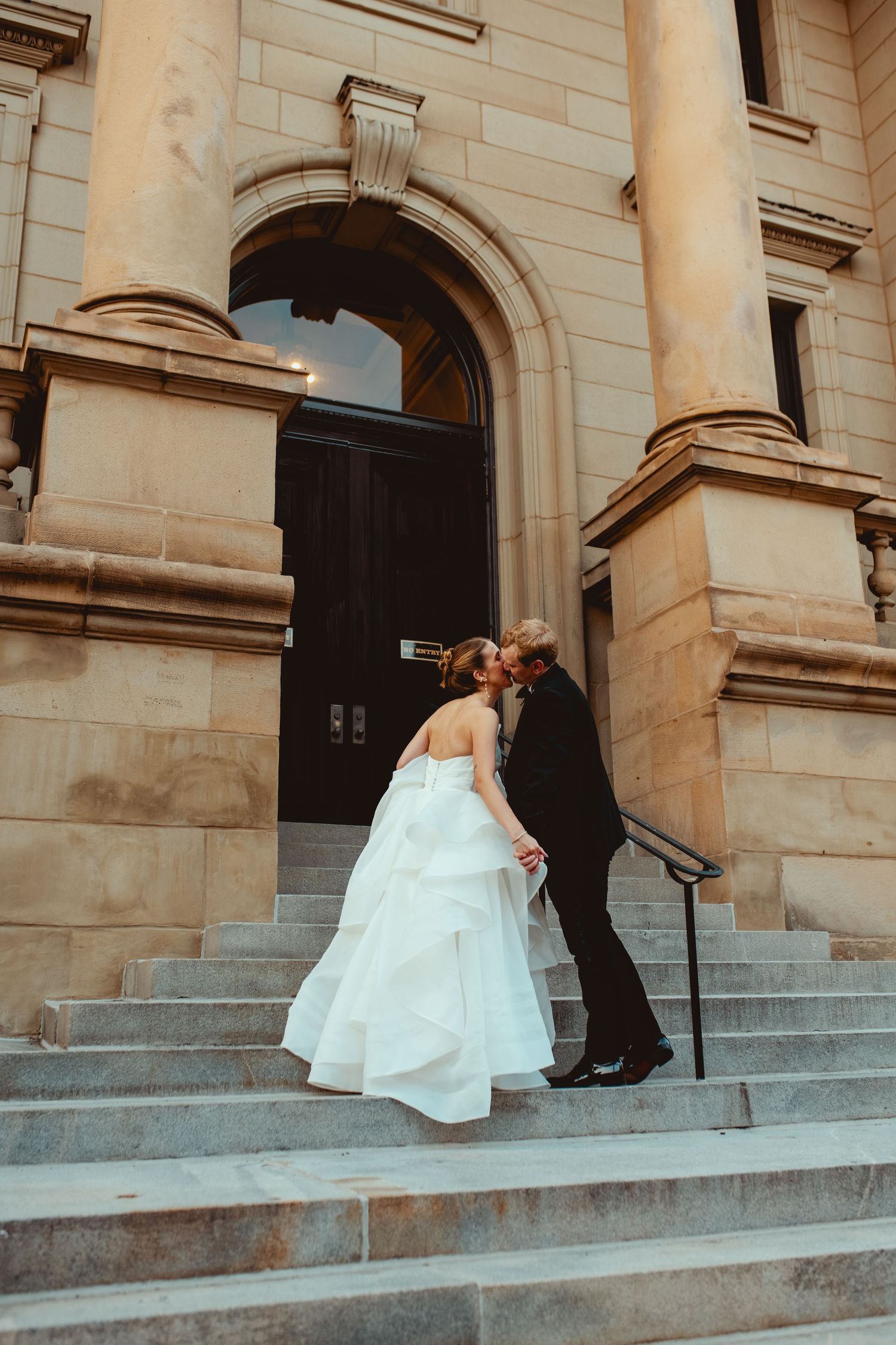 A couple poses dramatically on stone steps of a grand historic building.