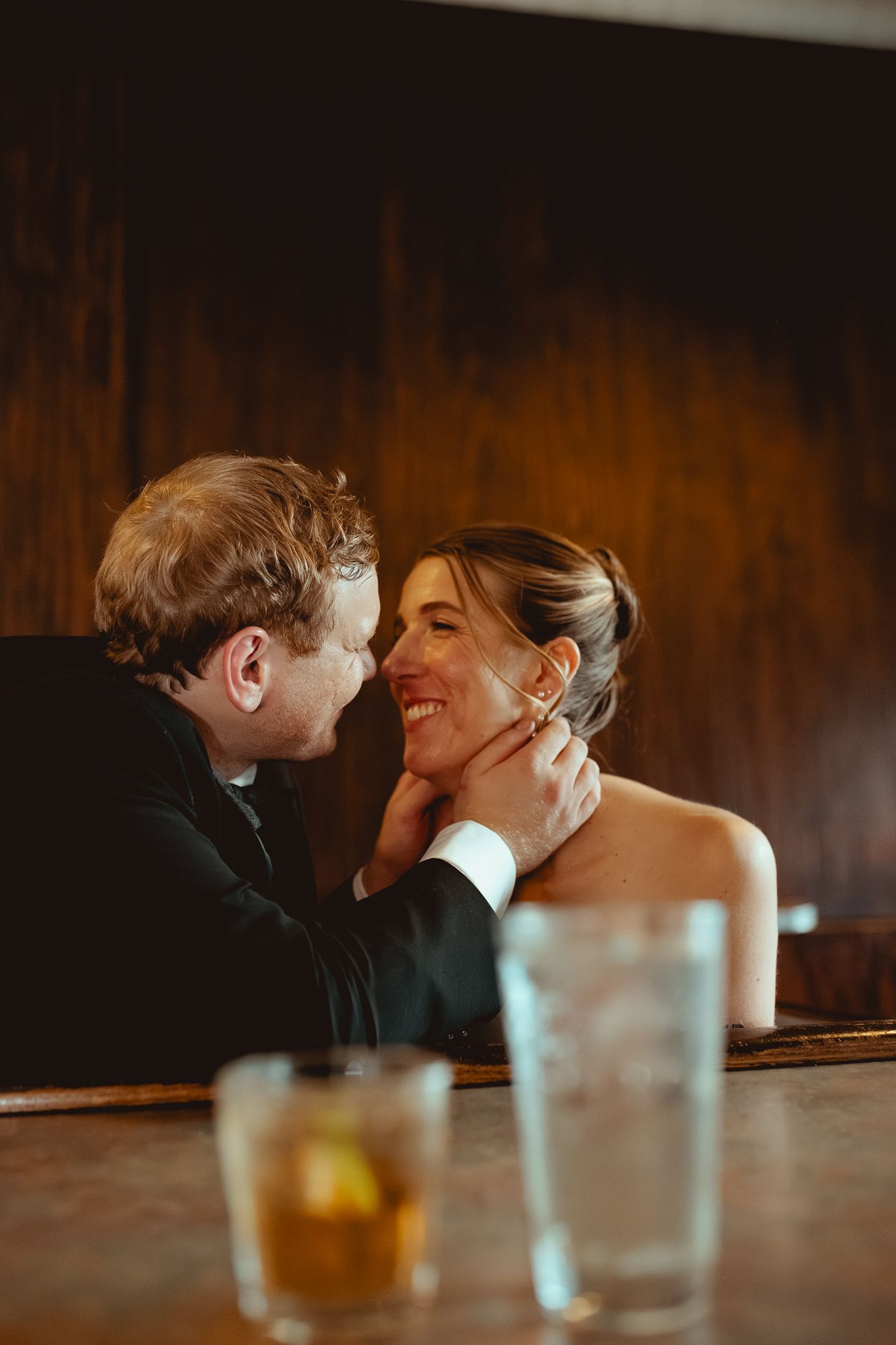 A couple shares an intimate moment at a dimly lit bar with drinks on the counter.