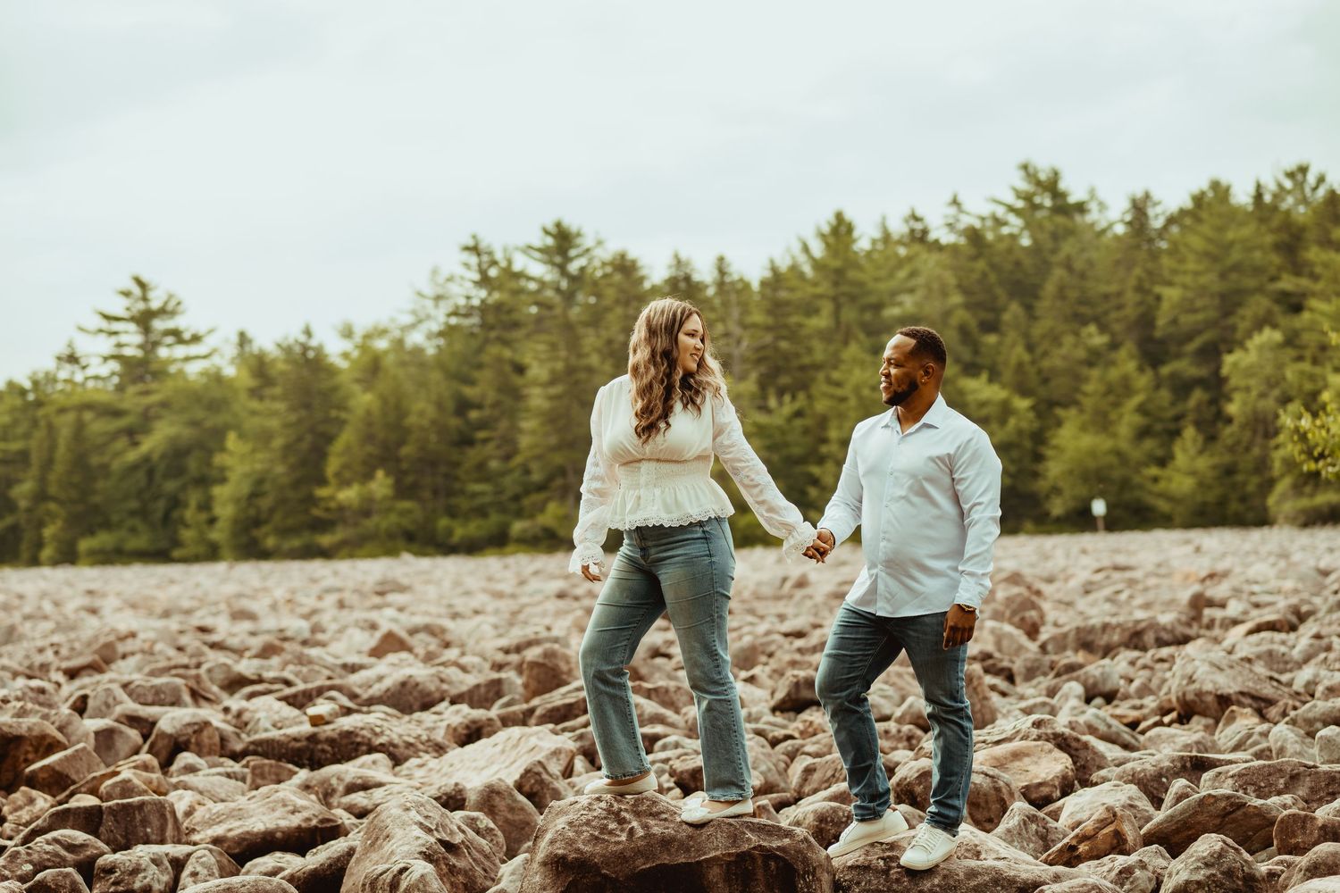 Couple holding hands and smiling while walking on rocky terrain with green trees in background.