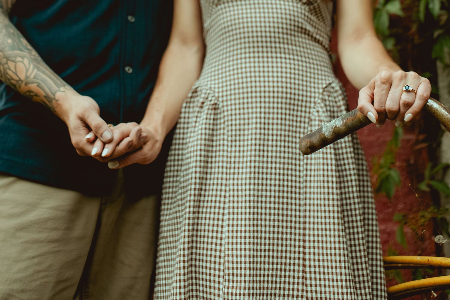 Couple holding hands, woman in plaid dress holding a cigar, man with tattoos in teal shirt, outdoor setting.