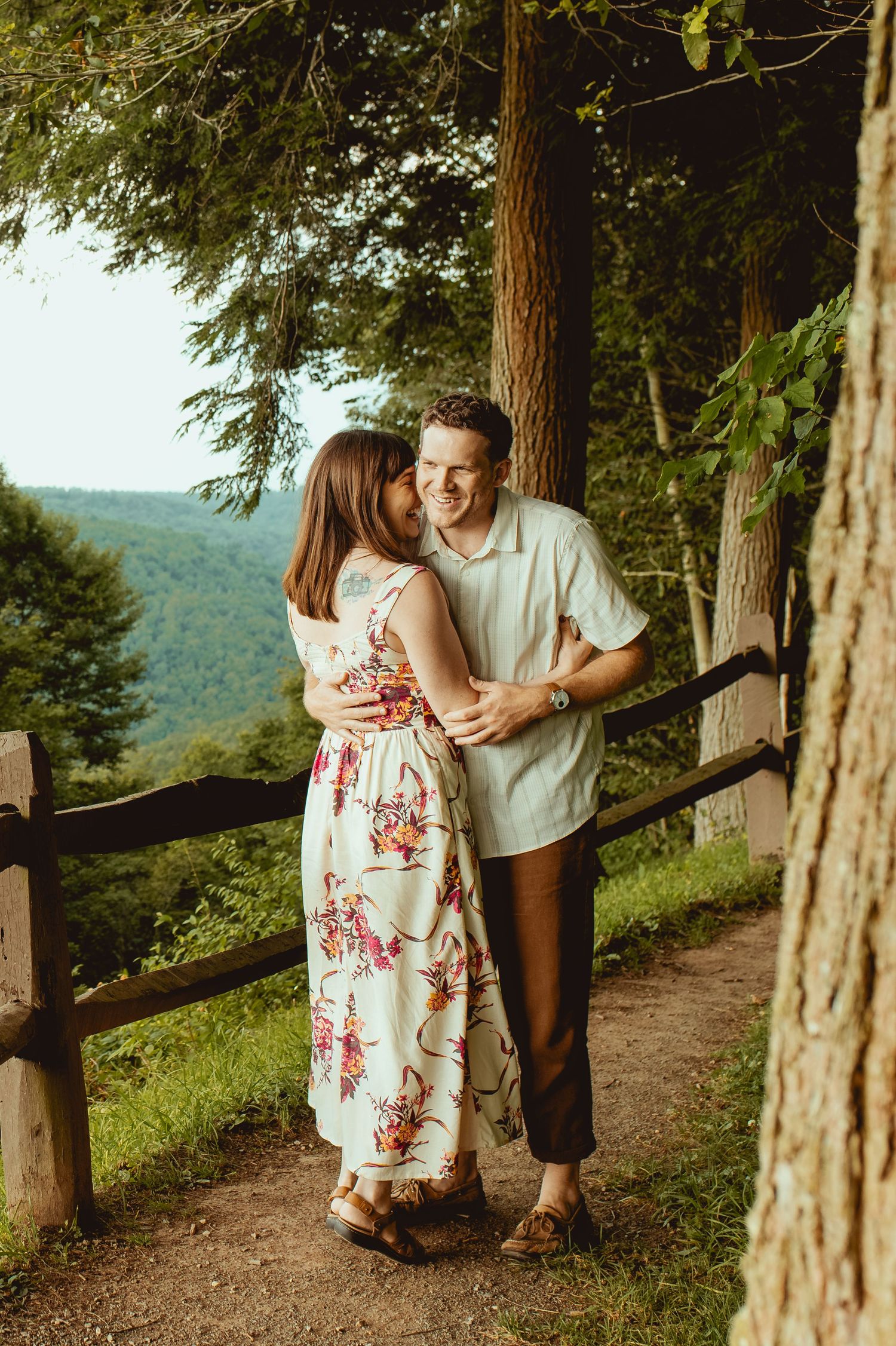 Couple embracing on a scenic wooded overlook path, woman in floral dress, man in light shirt, lush green hills behind them.