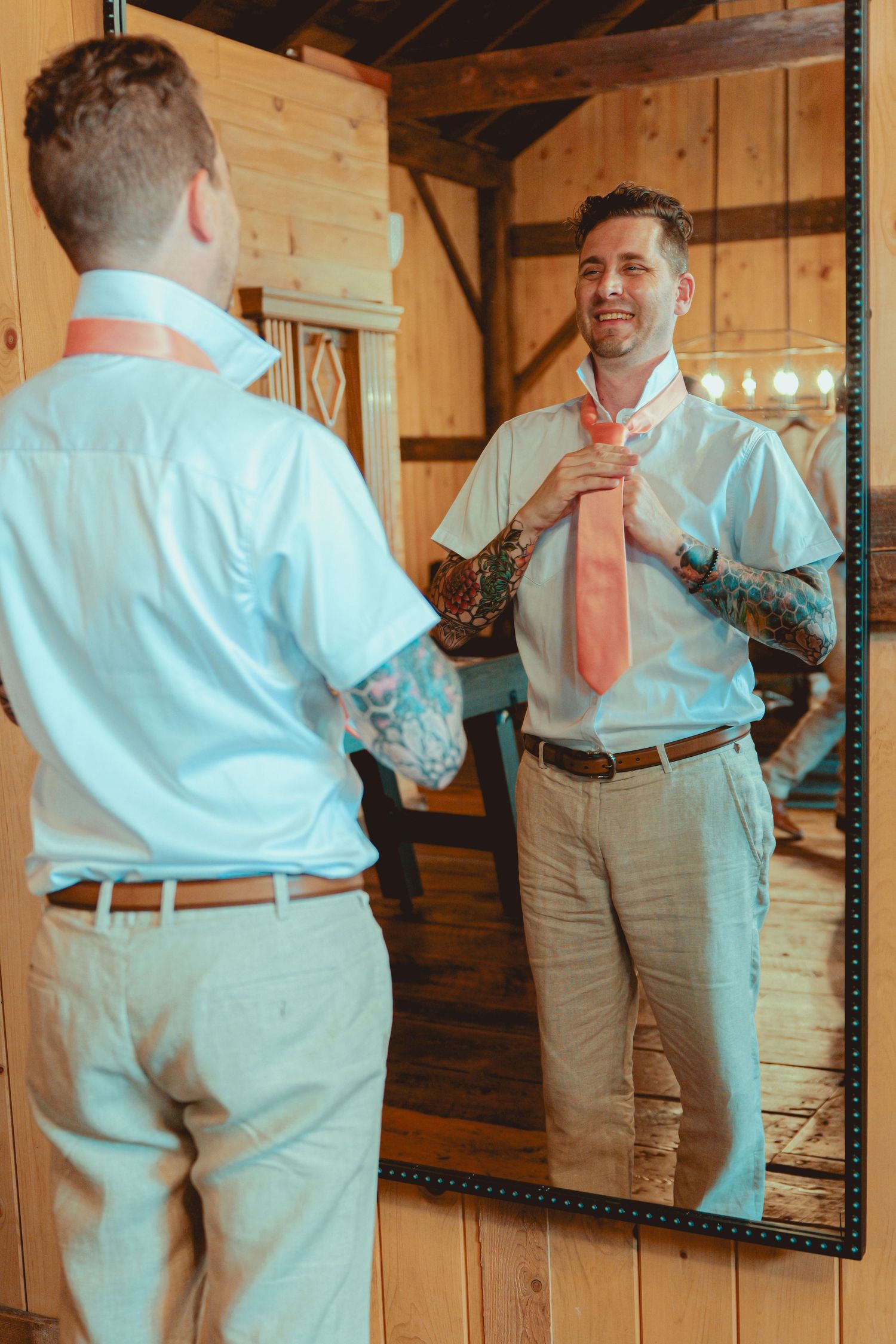Tattooed groom adjusting coral tie in mirror, wearing light blue shirt and khaki pants in rustic barn.