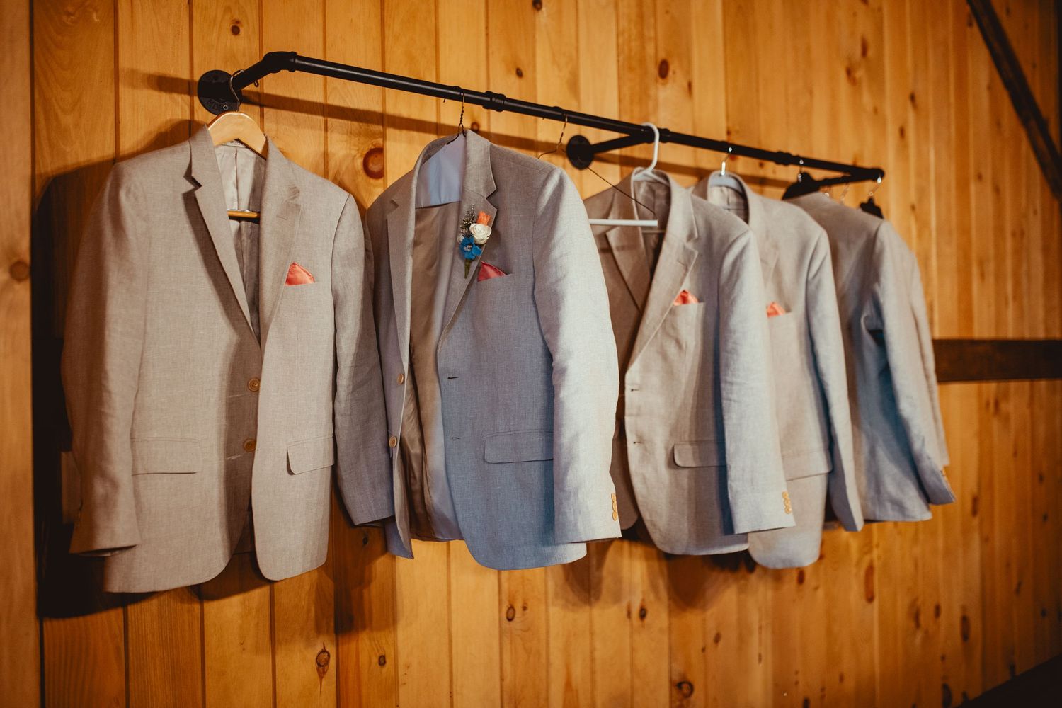 Four groomsmen suits in shades of gray and beige hang on a rustic wooden wall rack with floral boutonnieres.