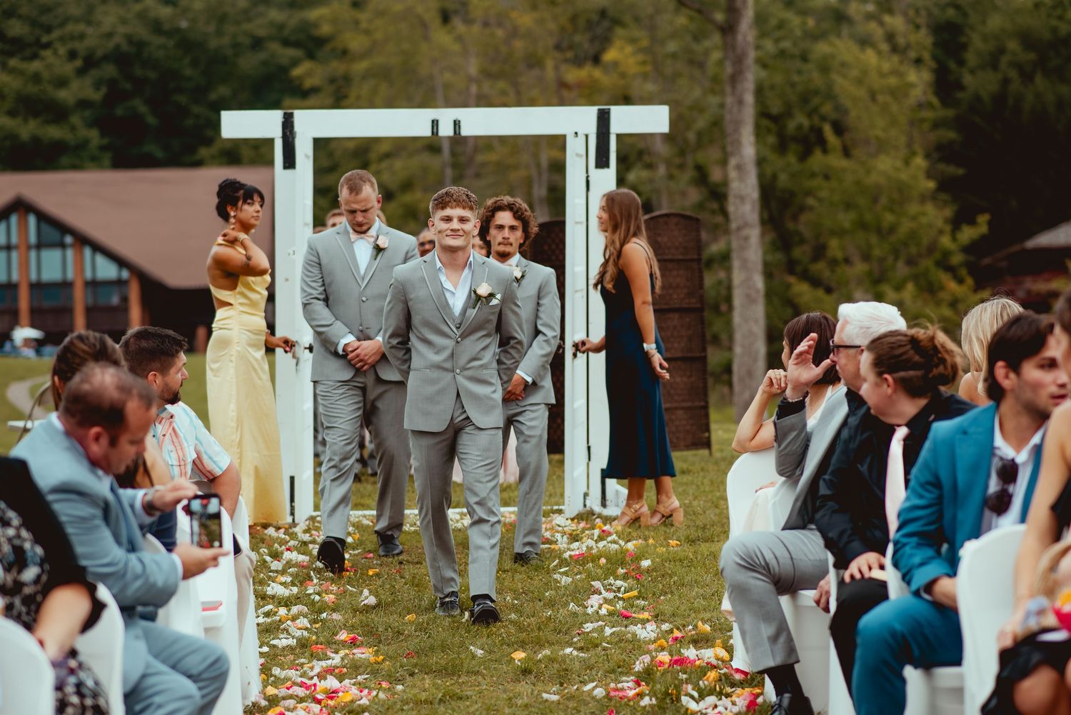 Two grooms walk down outdoor wedding aisle in gray suits, guests seated on sides, floral petals on grass.