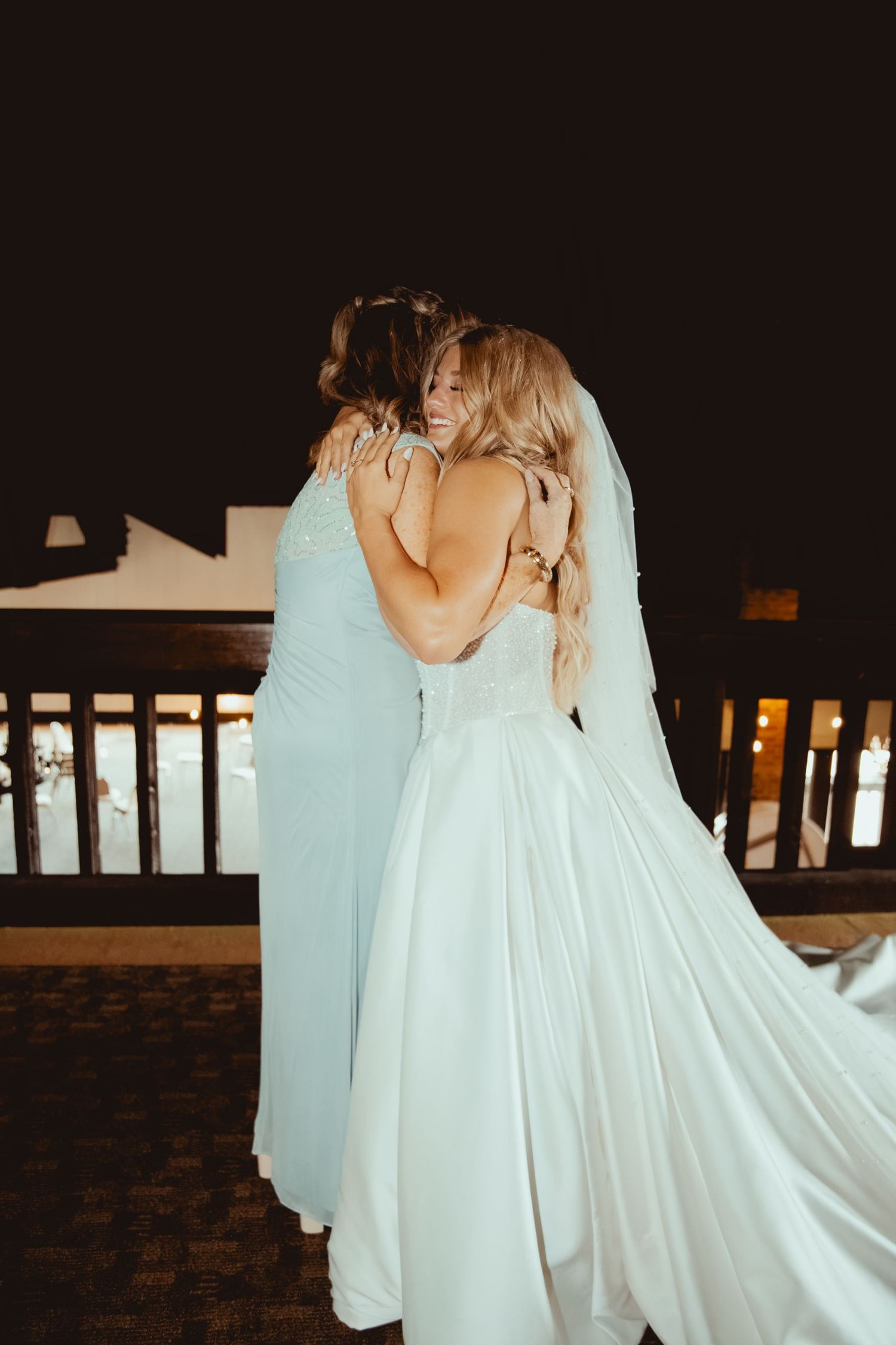 Bride and bridesmaid share an emotional hug on a balcony during wedding night celebrations.