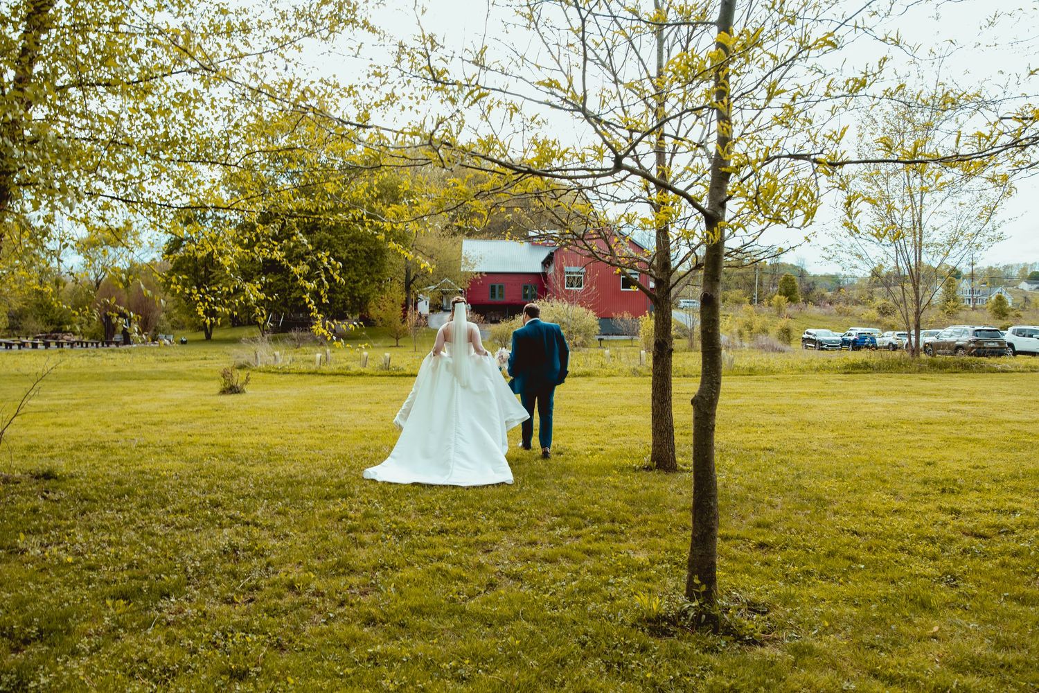A bride and groom walk hand in hand across a green lawn toward a red barn on their wedding day.