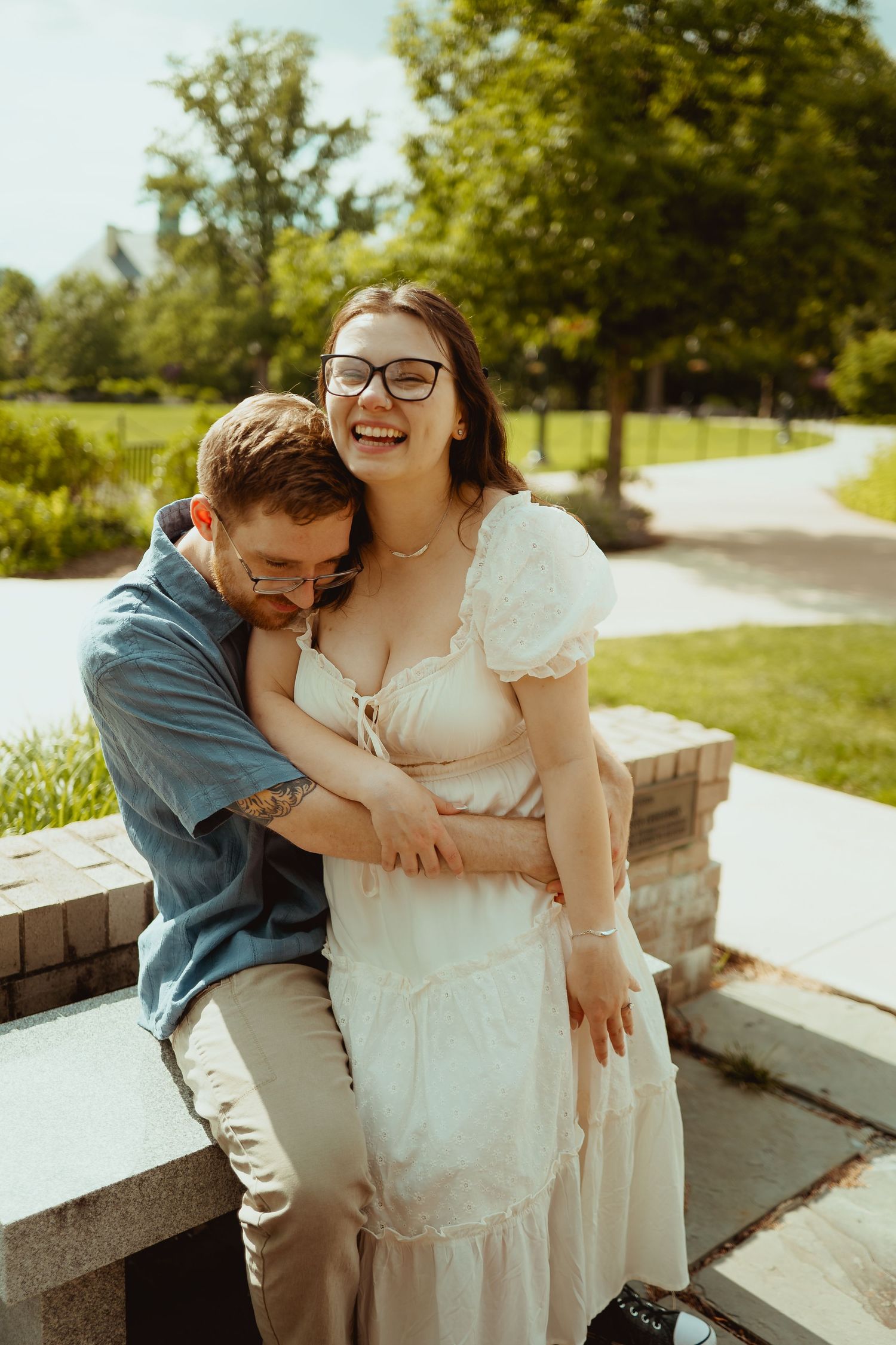 Couple laughing and embracing outdoors in a sunny park, woman in white dress with glasses, man in blue shirt.