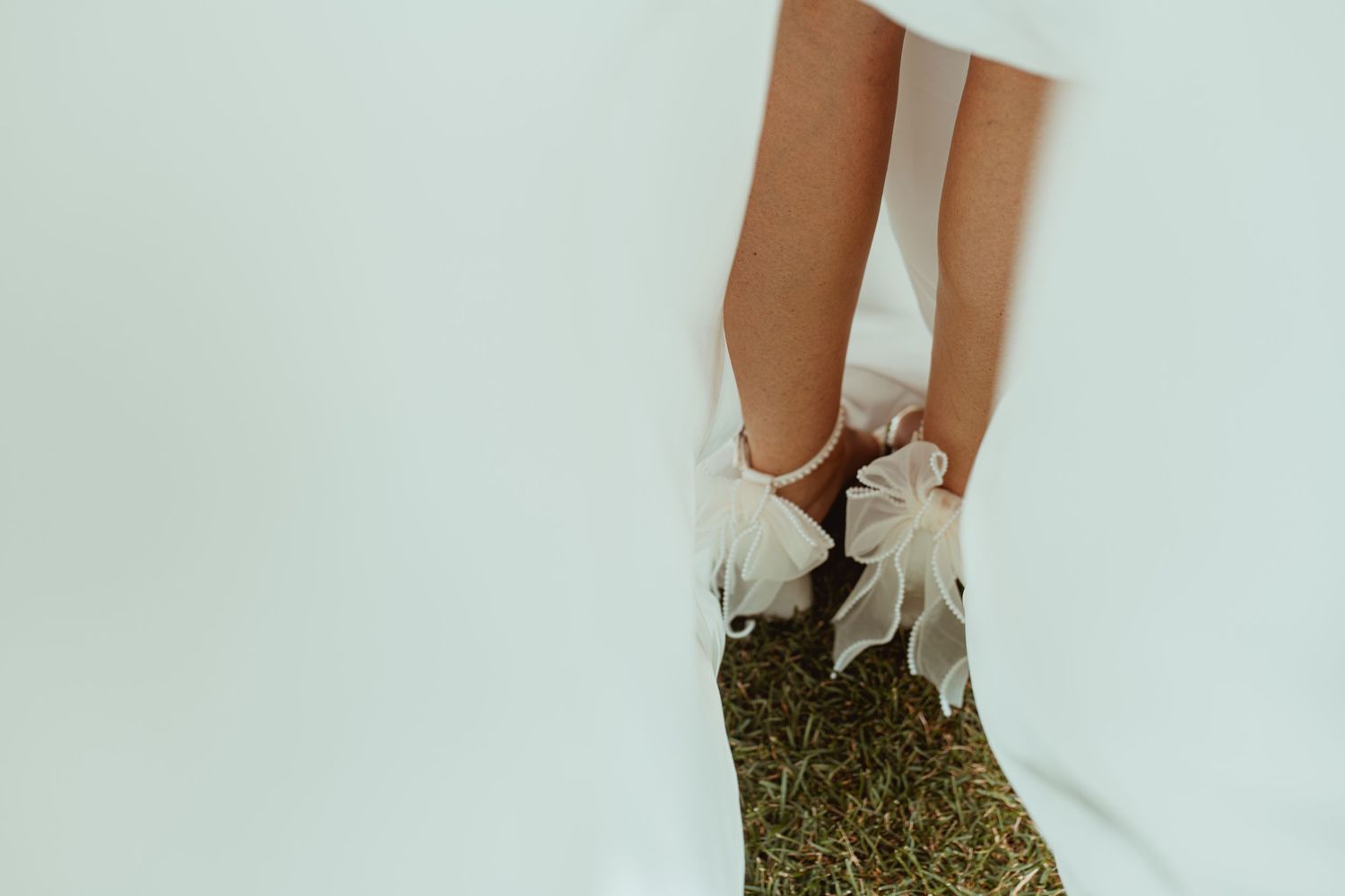 Bride wearing white wedding dress with elegant bow-adorned heels standing on green grass.