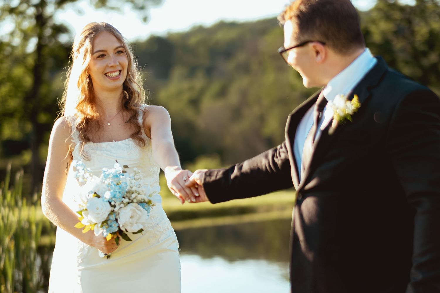 Bride and groom holding hands outdoors near a lake, bride smiling and holding a white and blue bouquet.