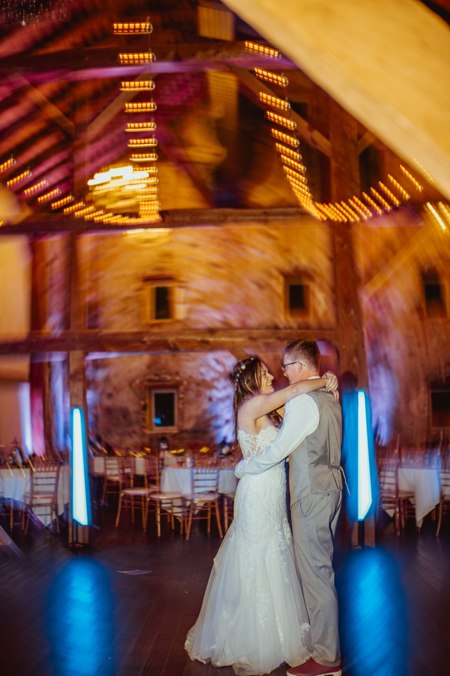 Couple dancing in a rustic wedding venue with blue uplighting and warm overhead lights creating a romantic atmosphere.
